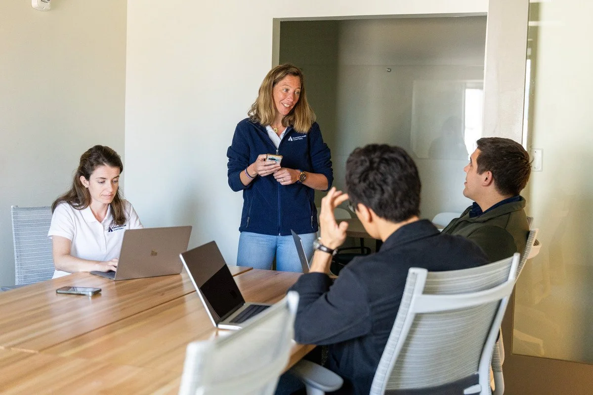 A woman standing at the head of a conference table, smiling and holding a smartphone, engaging with three seated young professionals working on laptops, in a modern office conference room.