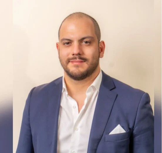 Hombre con barba y cabello rapado, usando traje azul y camisa blanca, posando frente a un fondo neutro.