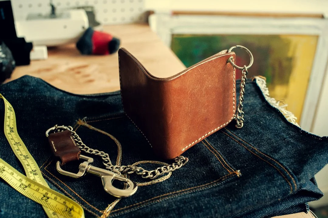 A leather keychain, a chain, and a leather wallet resting on a pair of folded denim jeans, with a yellow measuring tape and a blurred background of a workshop or studio.