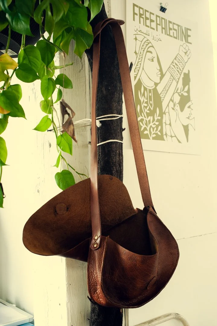A brown leather bag hanging from a nail or hook on a white-painted wooden post, with green leaves and a poster in the background.