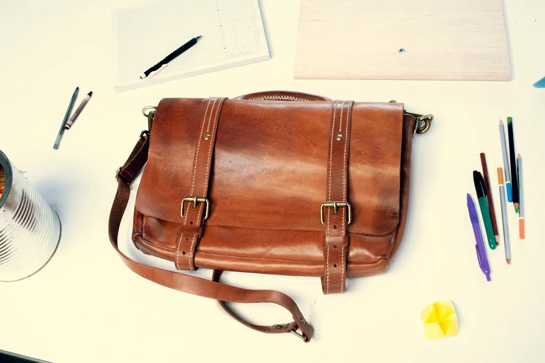 Top view of a work desk with a brown leather messenger bag in the center, surrounded by pens, a notepad, a wooden board, a yellow paper swan, and a silver can of snacks.