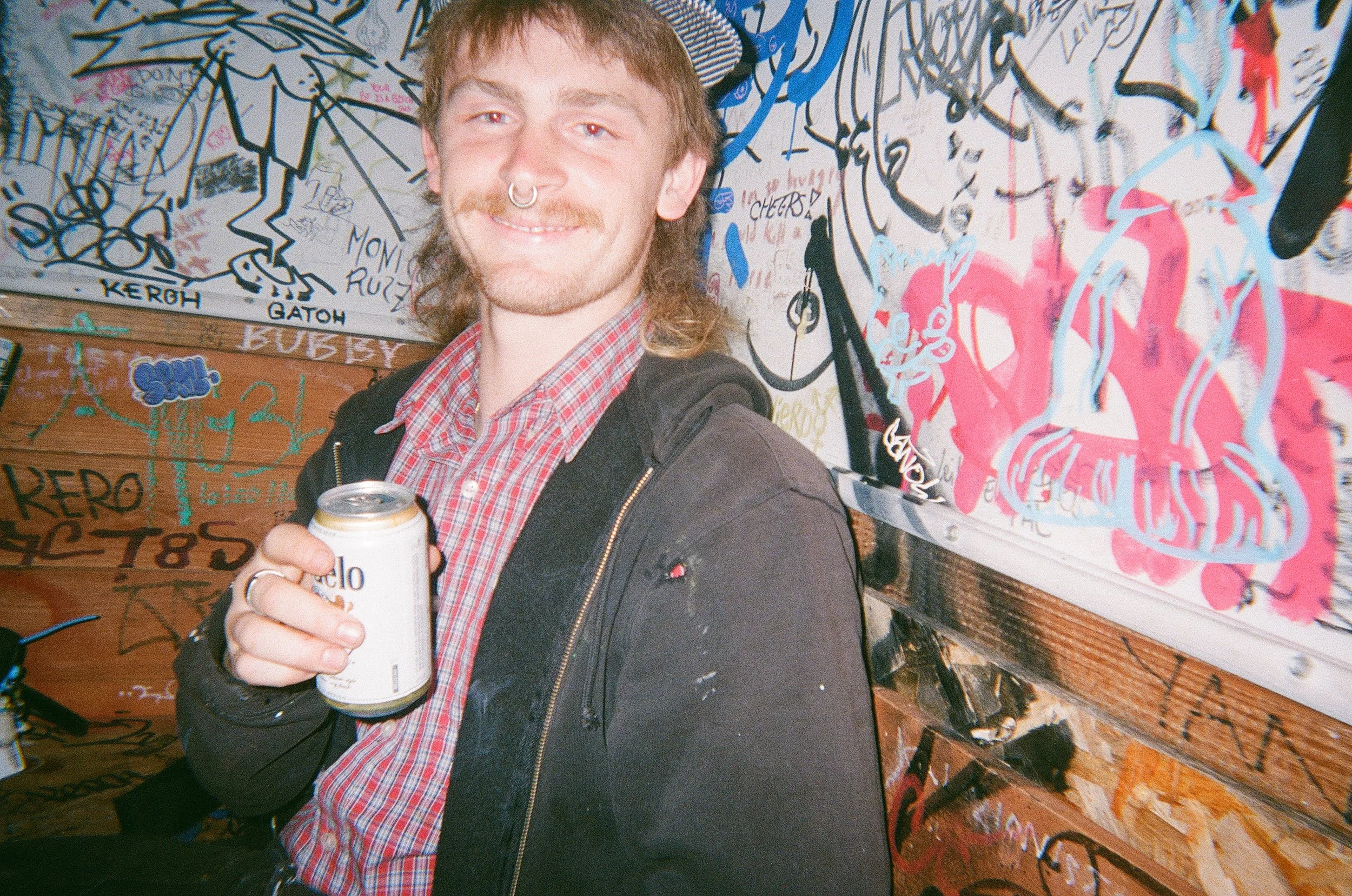A smiling young man with a mustache and long hair, wearing a nose ring, a red plaid shirt, and a black jacket, holding a can of beer, standing in front of a graffiti-covered wall.