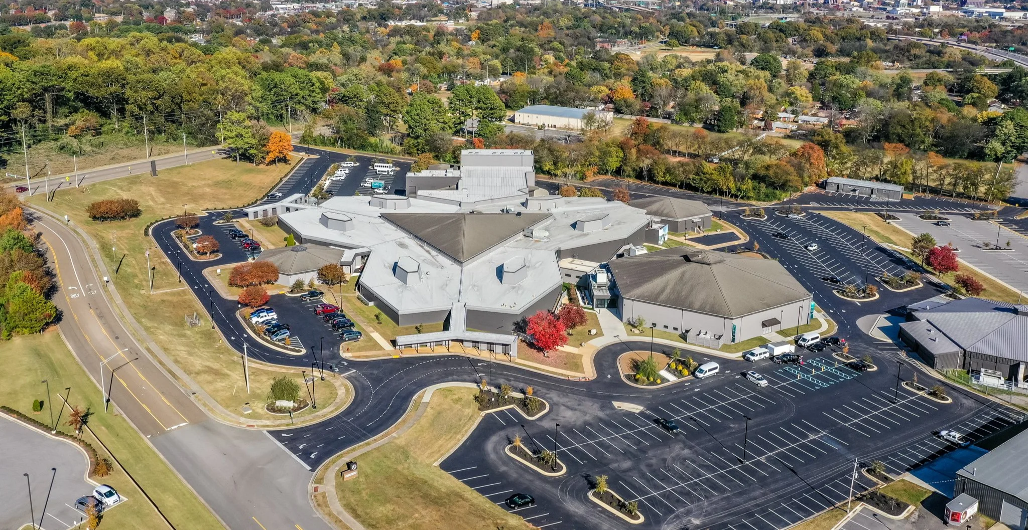 Aerial view the Rock Family Worship Center - a church in Huntsville Alabama surrounded by a city with trees, buildings, parking lots, and roads, set against a backdrop of distant hills and a clear blue sky.