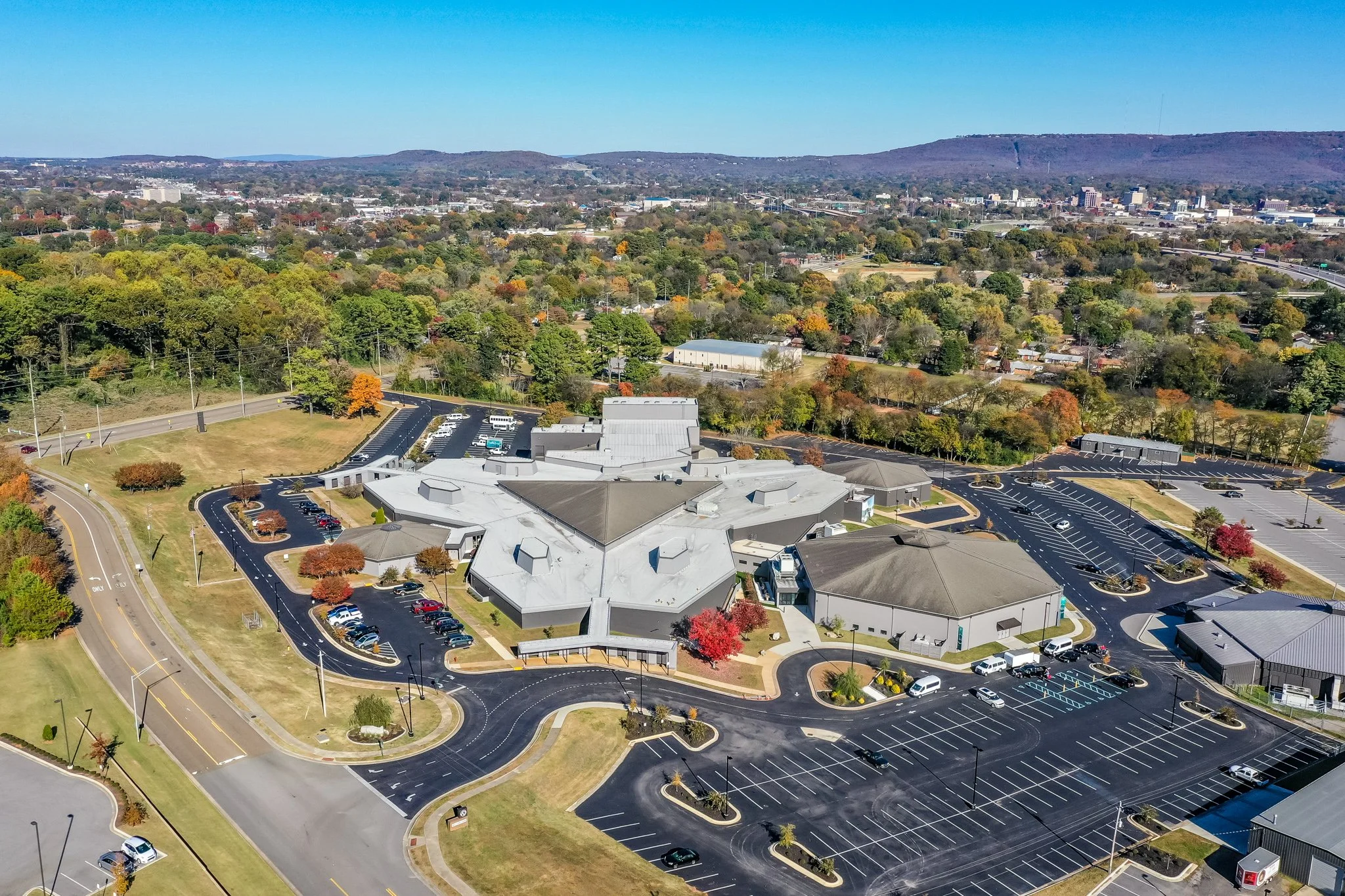 Aerial view the Rock Family Worship Center - a church in Huntsville Alabama surrounded by a city with trees, buildings, parking lots, and roads, set against a backdrop of distant hills and a clear blue sky.