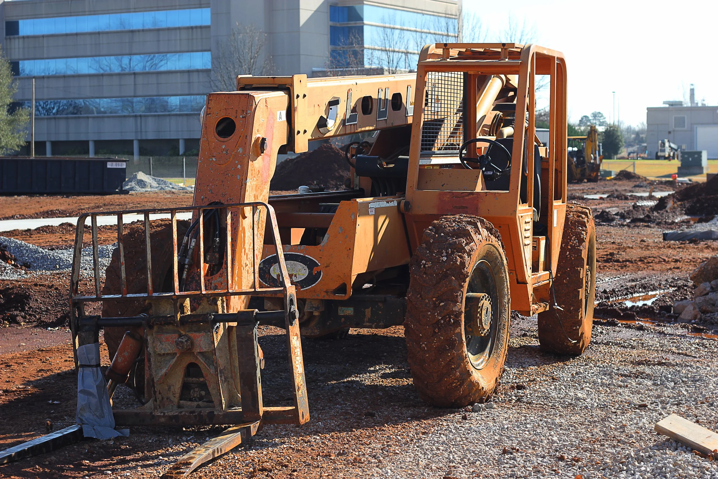 Construction site with orange heavy machinery, including a bulldozer and excavator, on muddy ground, with modern glass buildings and leafless trees in the background.