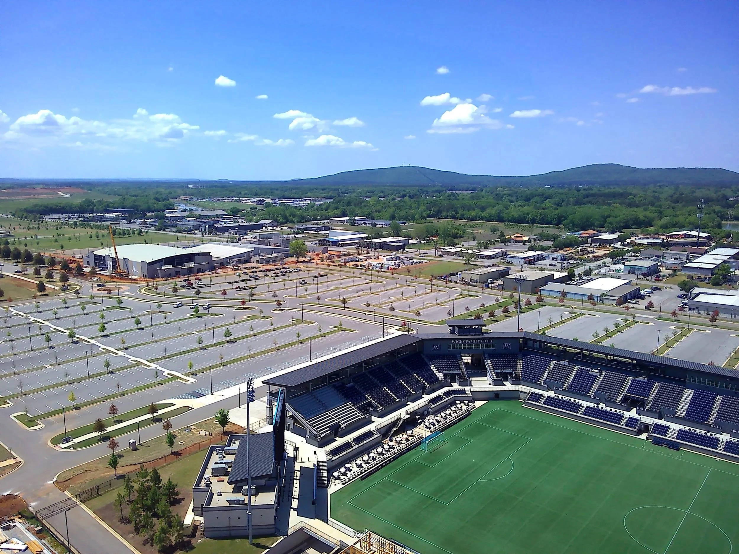 Aerial view of a Wicks Family Field at Joe Davis Stadium with a green field and seating area, surrounded by a parking lot, buildings, and green landscape with hills under a blue sky with clouds.