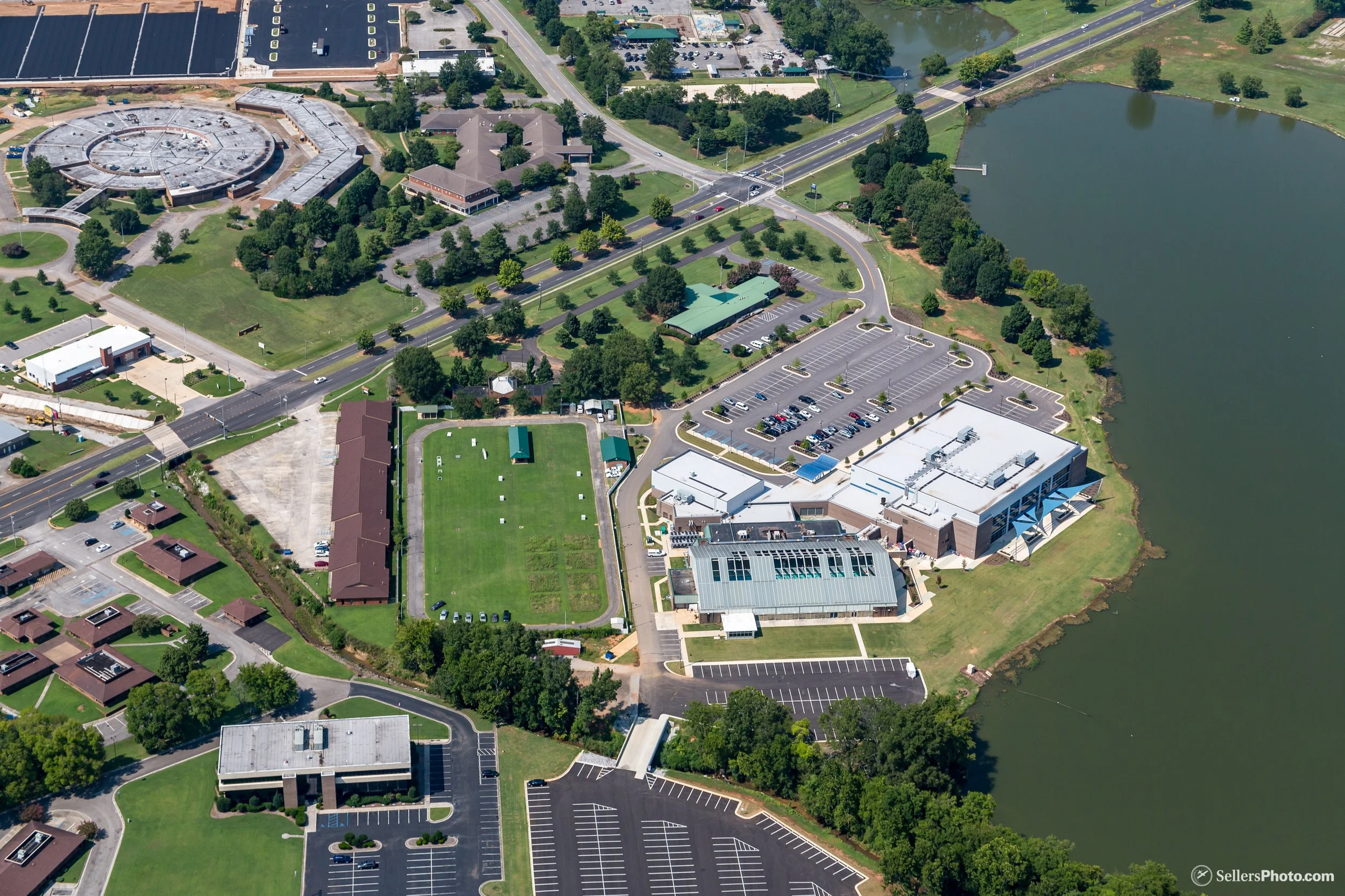 Huntsville Aquatic Center. An aerial view of a suburban area including parking lots, a large circular building, green spaces, a pond, and roads.
