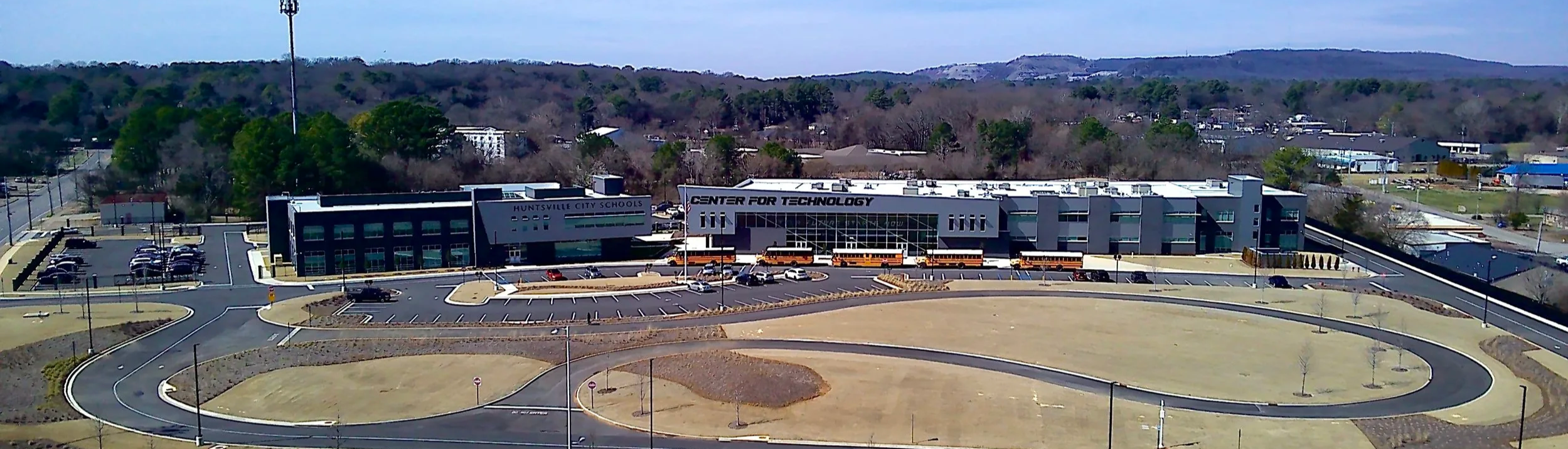 Aerial view of a modern school building labeled 'Huntsville City Schools Center for Technology,' with a parking lot, bus circle, and surrounding roads, trees, and hills in the background.