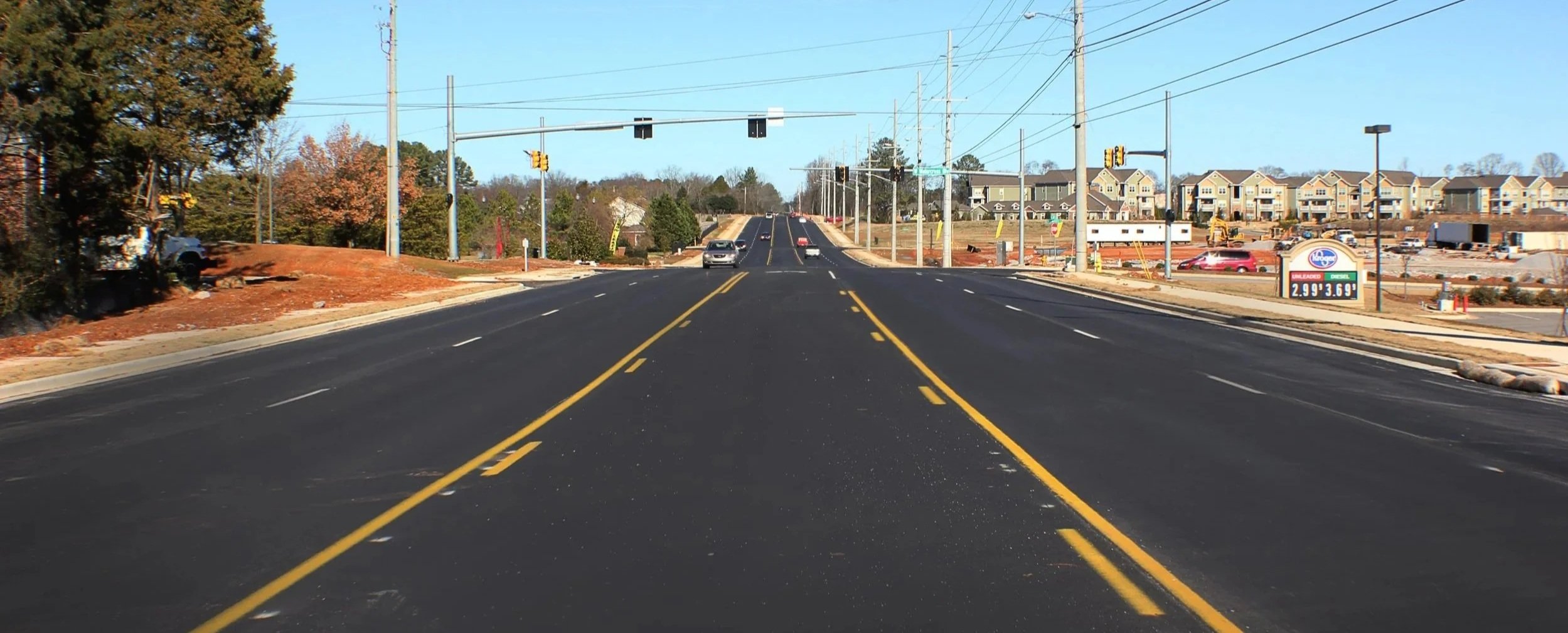 A newly paved road with yellow dividing lines, cars driving, traffic lights, and construction on the right side with residential buildings in the background.