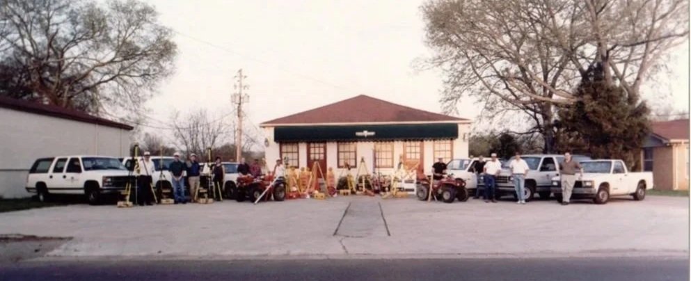 People gathered outside a building with a prominent green awning surrounded by several parked white vehicles and a bunch of surveying equipment