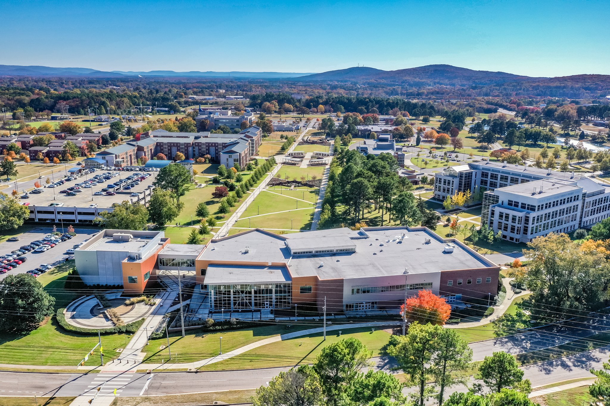 Aerial view of the University of Alabama in Huntsville college campus with buildings, parking lots, green lawns, trees, a pond, and surrounding hills in the background during autumn. This depicts the walkway corridor Johnson & Associates designed.
