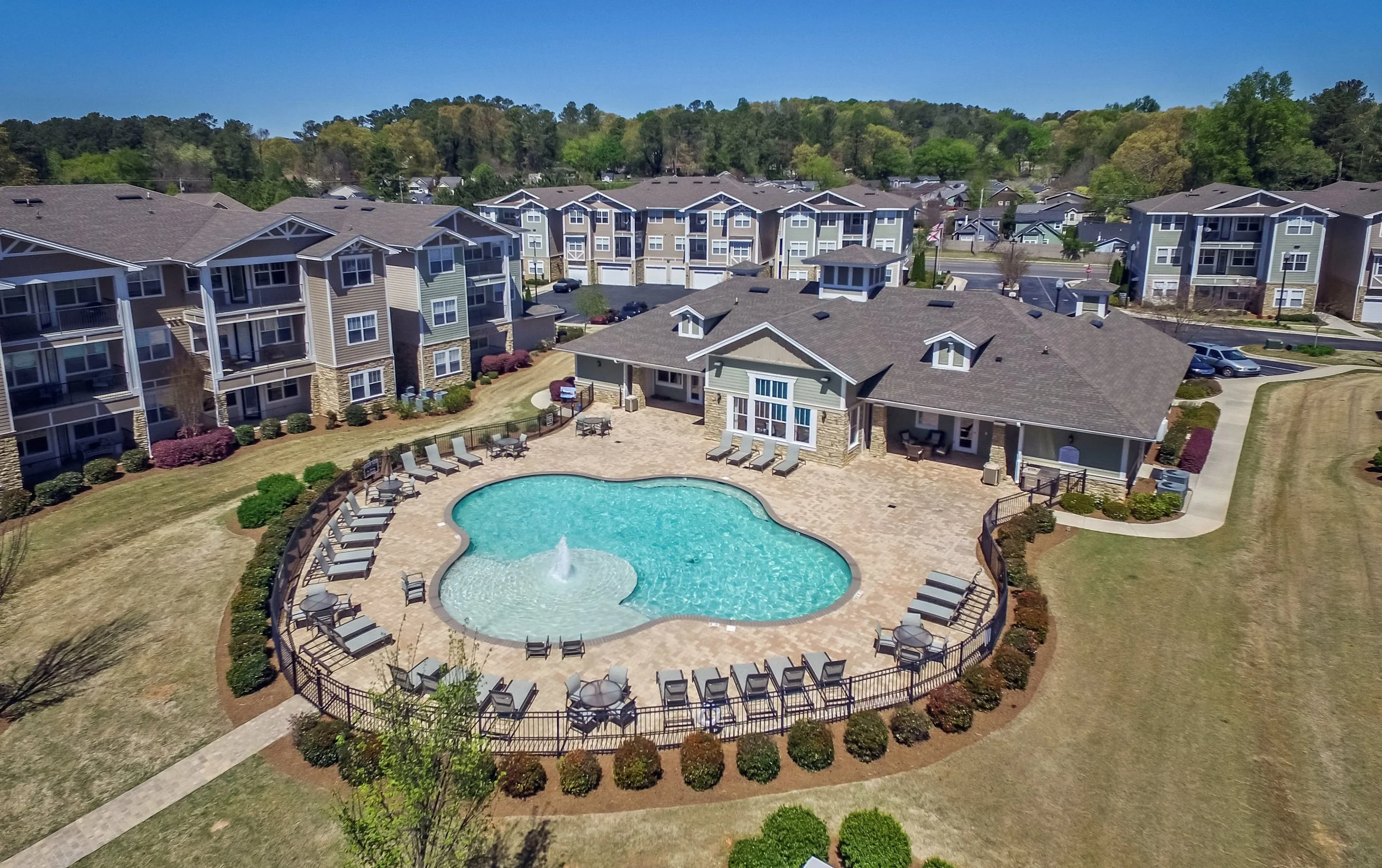 Aerial view of an apartment complex with buildings, a swimming pool, poolside lounge chairs, and surrounding greenery.