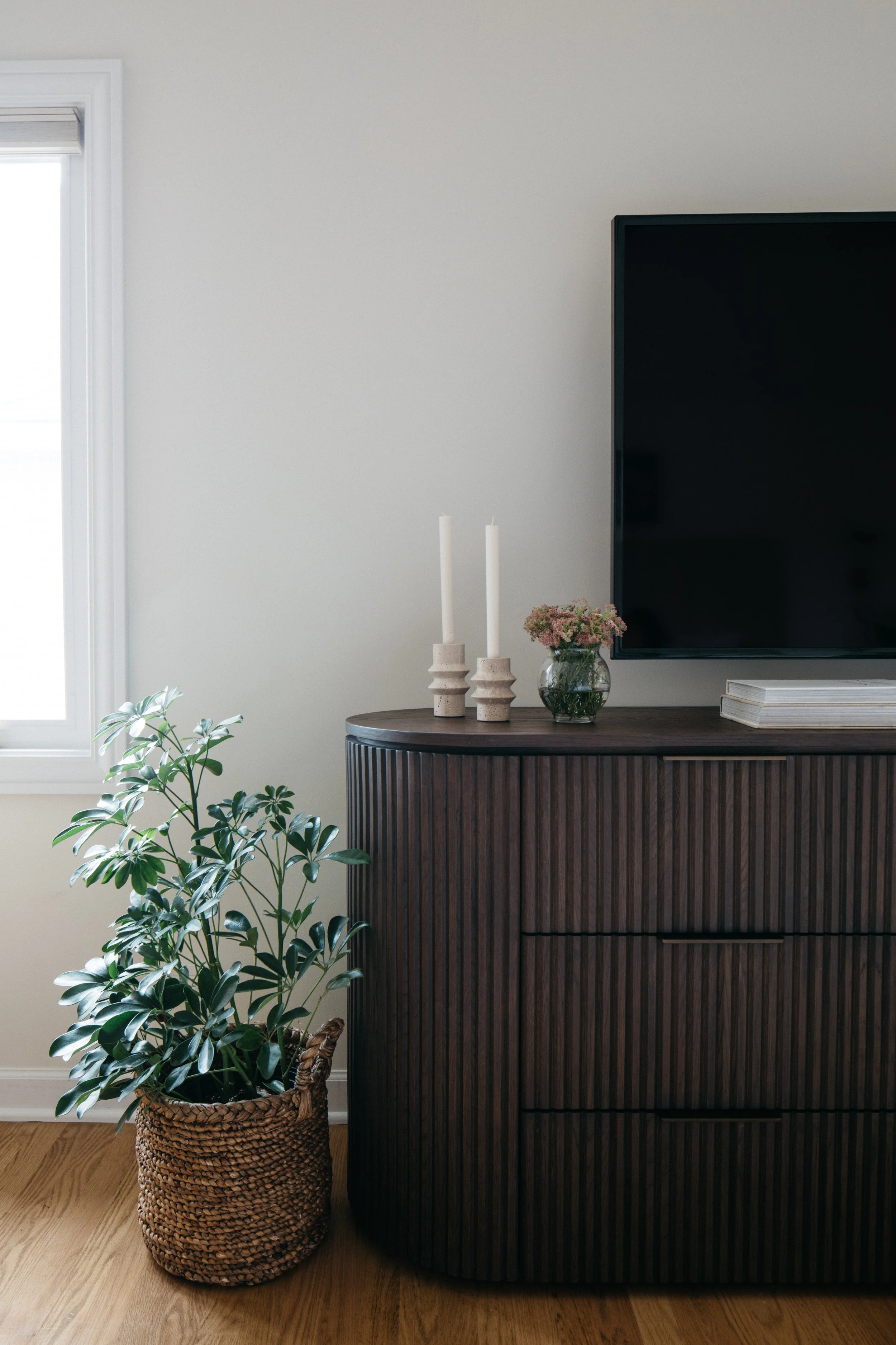 Living room corner with a wicker basket holding a green leafy plant, a dark wooden sideboard, two white candles in beige holders, a glass vase with pink flowers, and a flat-screen TV mounted on the wall.