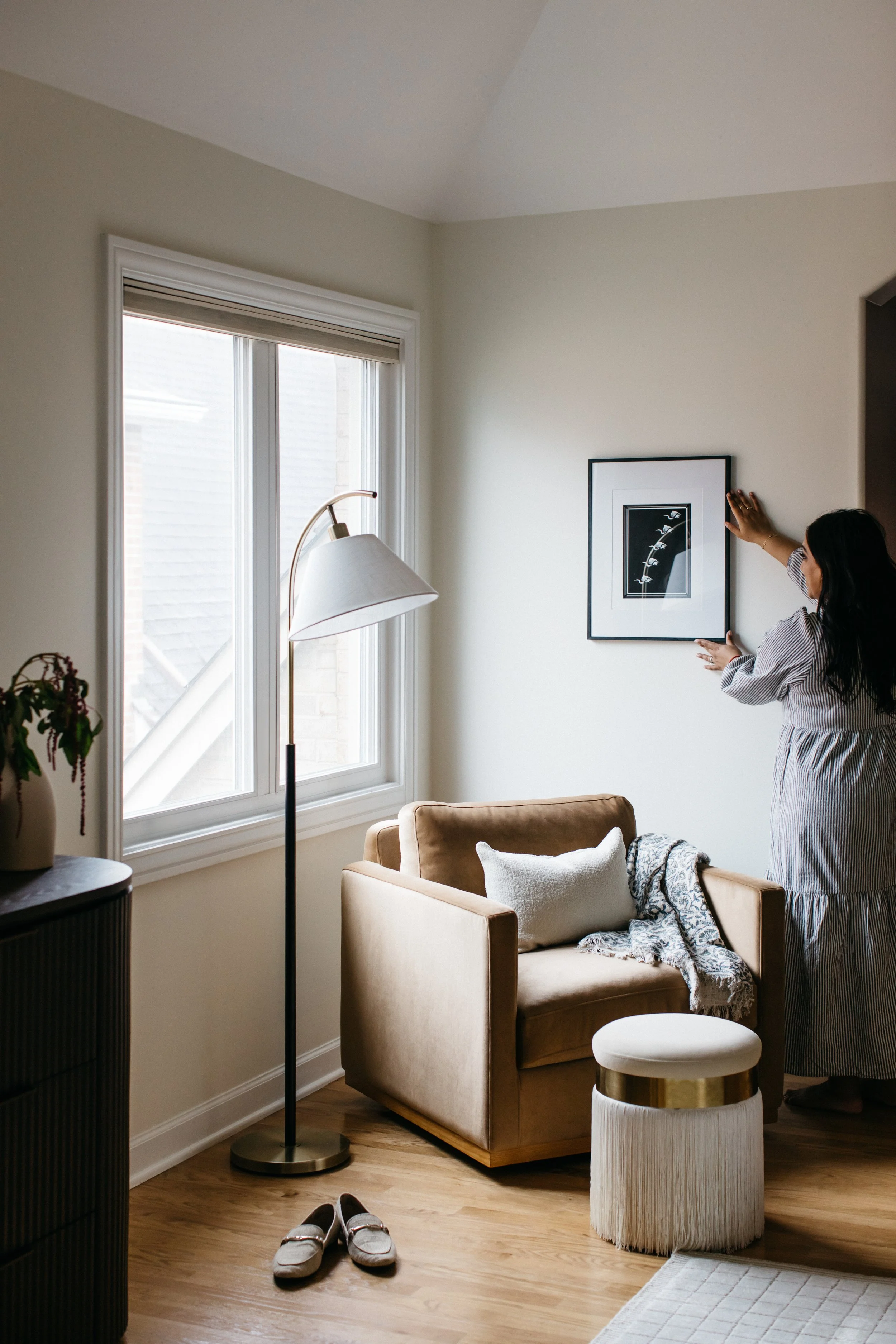 Woman hanging a framed picture on a white wall in a cozy living room with a beige armchair, floor lamp, and pair of slippers on the wooden floor.