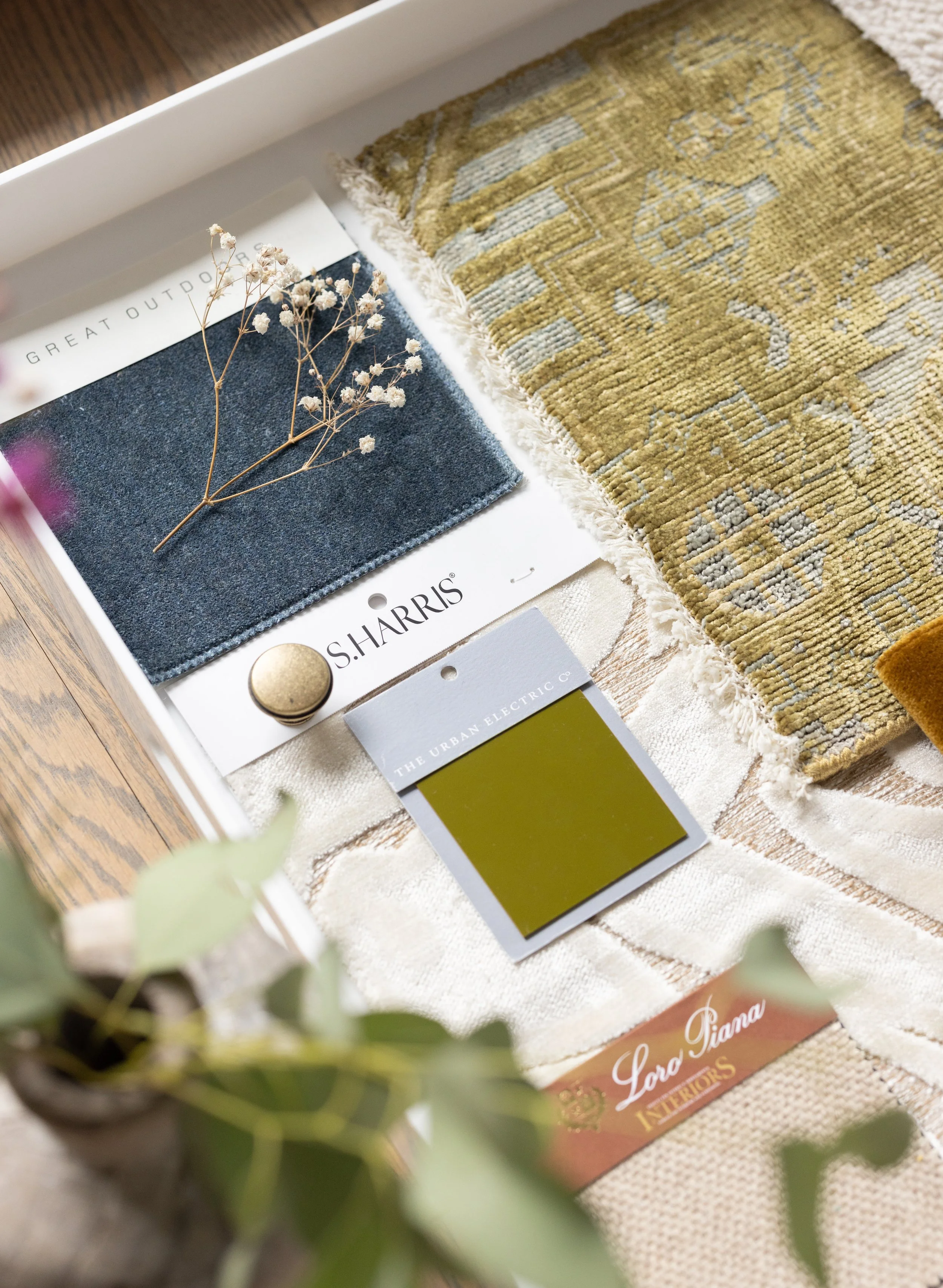 Interior design color and fabric samples on a white tray, including a dark blue fabric swatch with a sprig of dried flowers, a beige and gold patterned rug, a green and yellow paint chip labeled 'The Urban Electric Co.', a brass round knob, and a label reading 'LORD PIANO INTERIORS', with a blurred plant in the foreground.