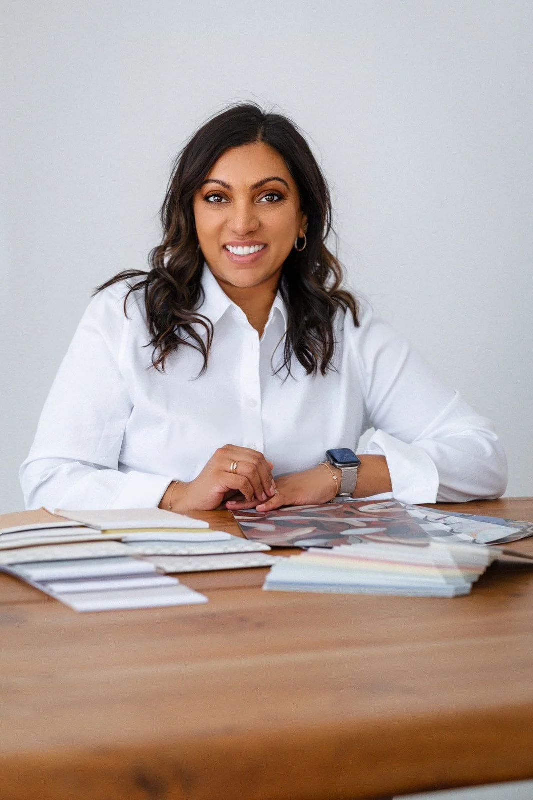 A woman with dark wavy hair, smiling, wearing a white shirt, sitting at a wooden table with color swatches and fabric samples, and a smartwatch on her wrist.