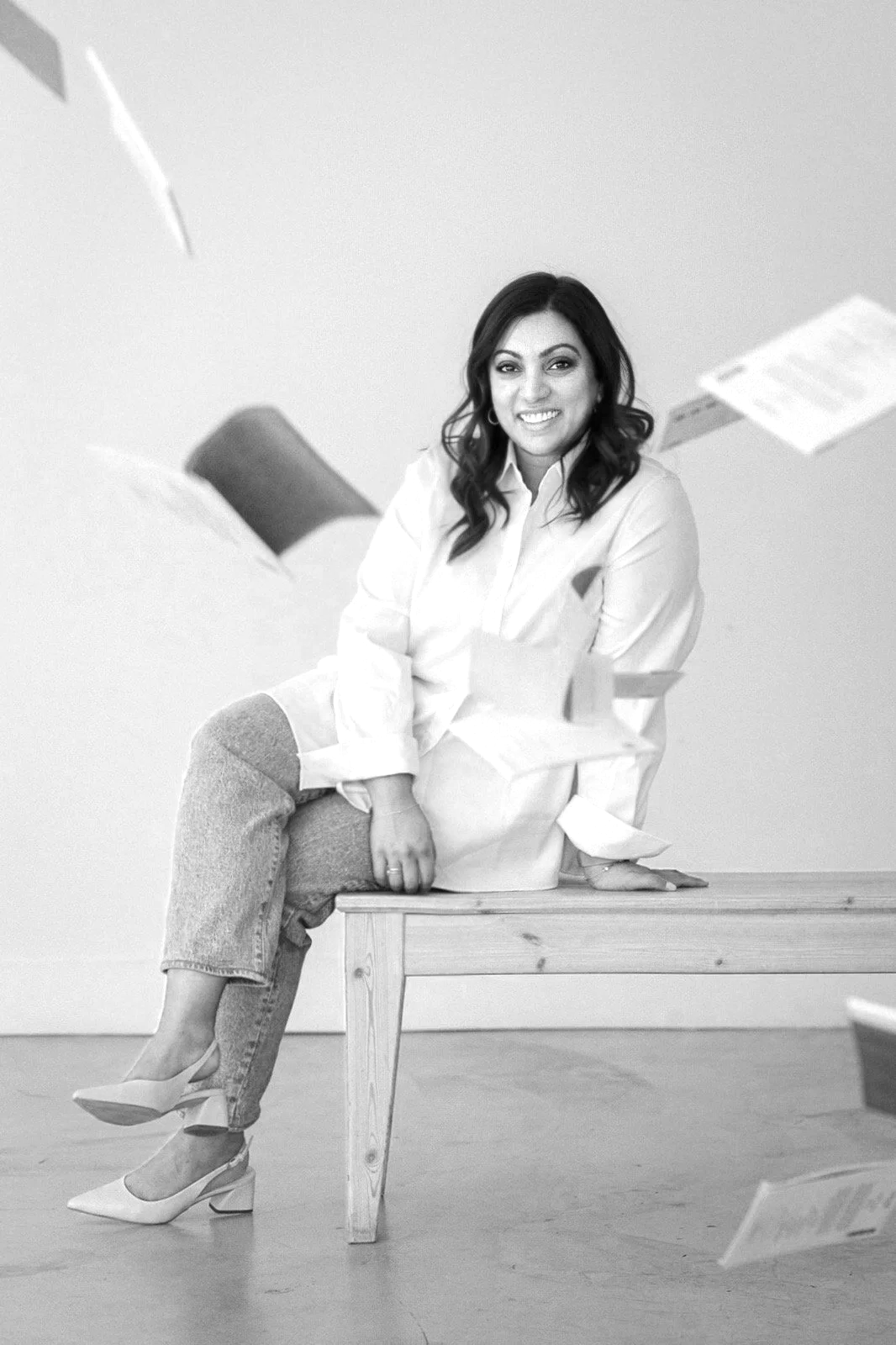 A woman with dark hair sitting on a wooden bench, smiling at the camera, with papers and folders flying around her in mid-air.