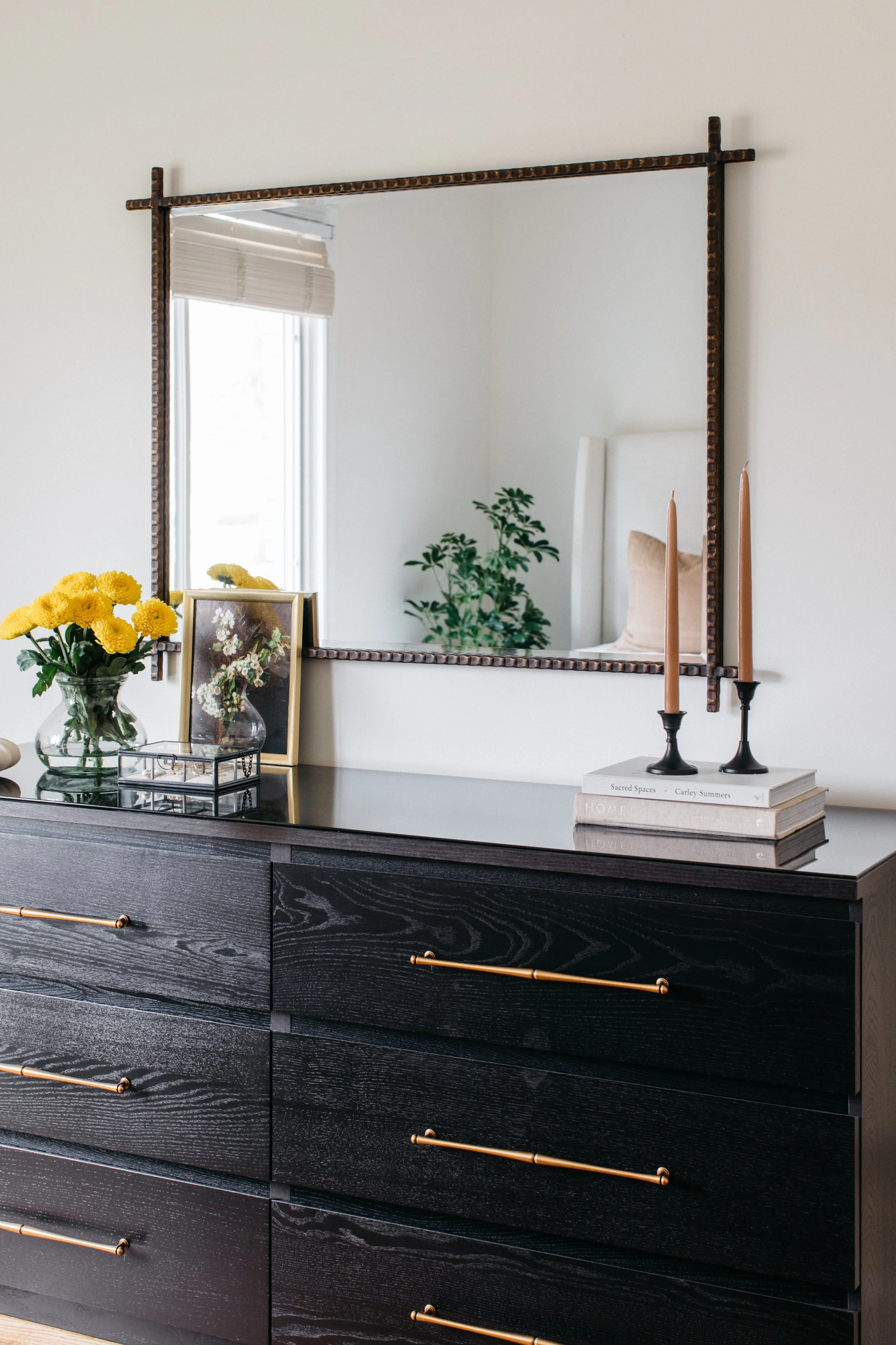 A black dresser with gold handles shown in front of a mirror on a white wall. The dresser is decorated with yellow flowers in a glass vase, a framed flower picture, two stacked books, and two tall candles in black holders.