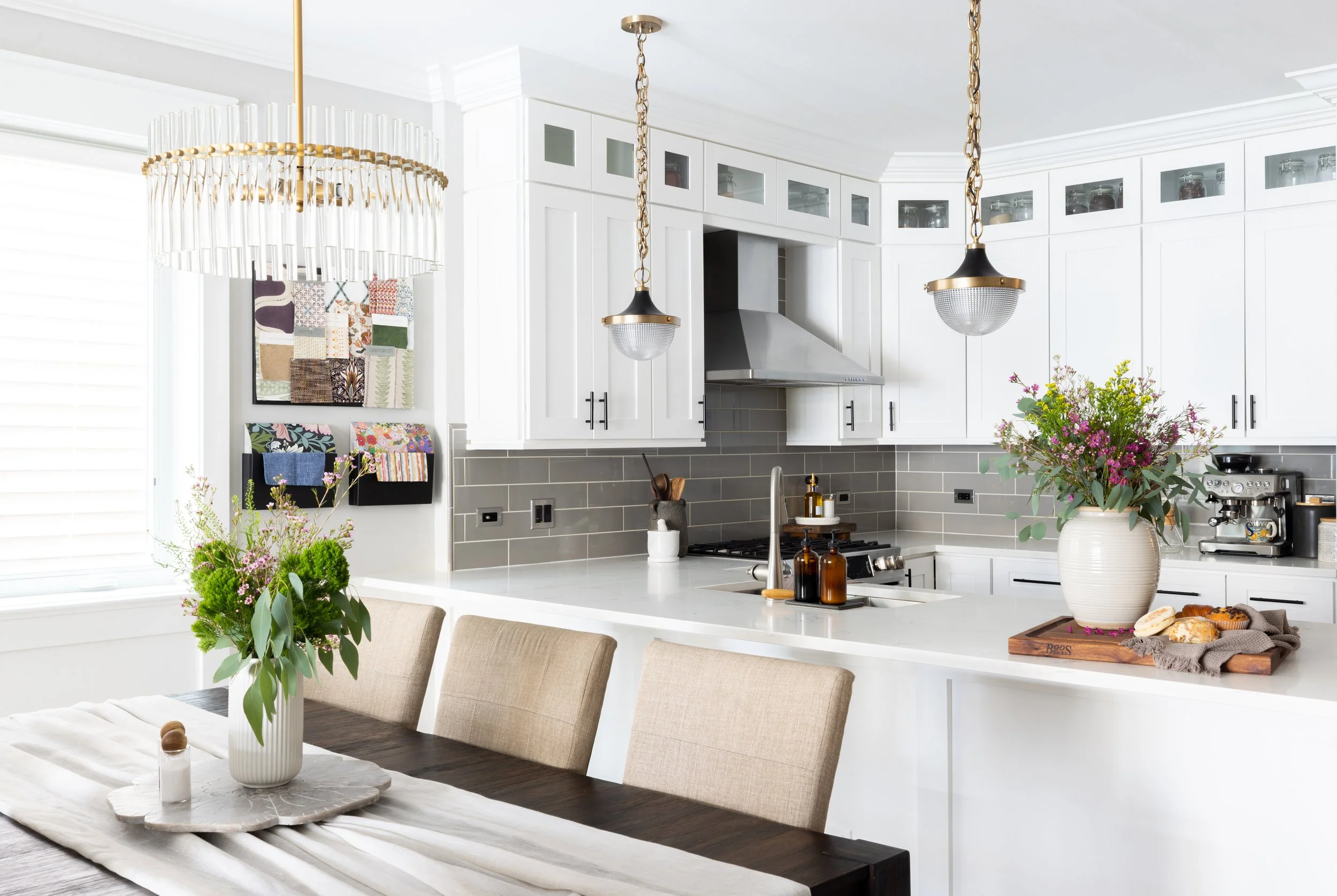 A bright, modern kitchen with white cabinets, gray tile backsplash, and a large white island counter. Two hanging pendant lights, a flower arrangement on the counter, and a vase with flowers on the dining table are visible.