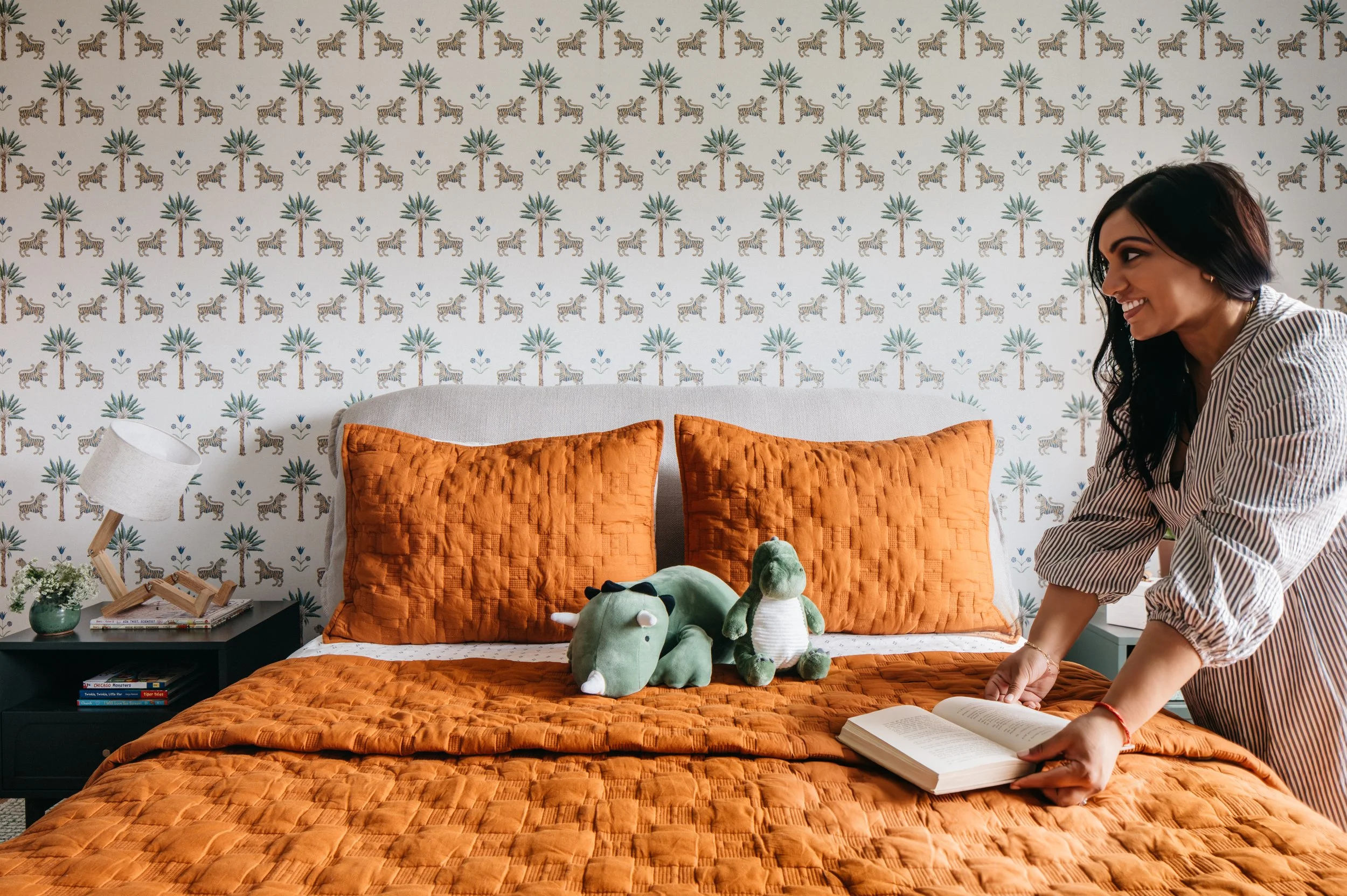 A woman is making a bed with orange bedding and two large matching pillows, one large stuffed hippo, and one stuffed dinosaur toy. There's a black nightstand with a small lamp, a small plant, and books on it. The wall has a palm and tiger wallpaper pattern.