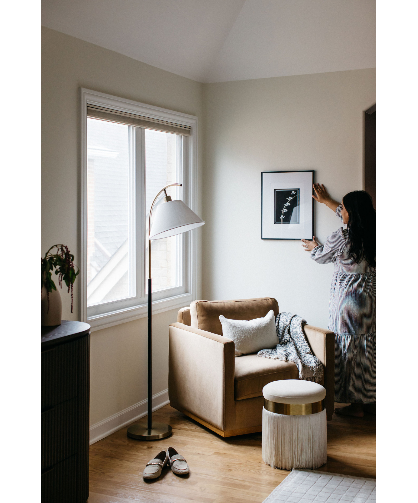 A woman hanging a framed picture on a light-colored wall in a cozy living room with a window, a standing lamp, a beige armchair, a white pillow, a patterned blanket, a small round ottoman, and a pair of slippers on the wooden floor.
