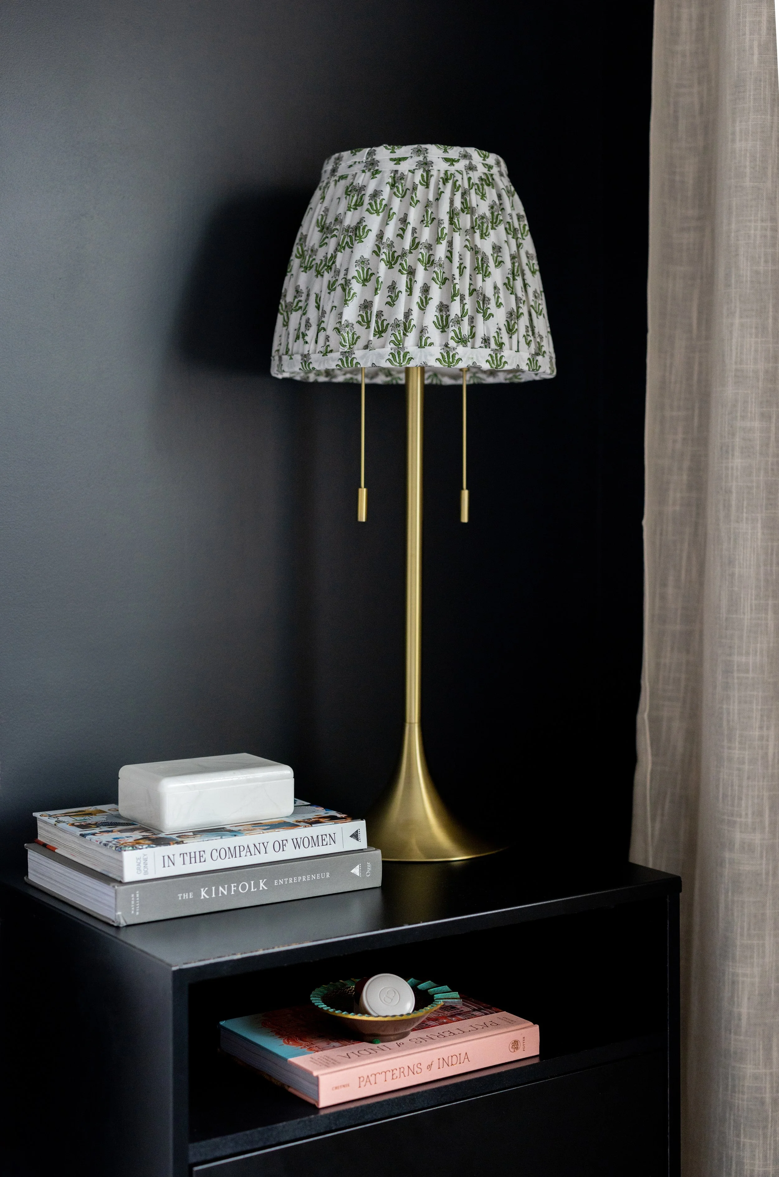 A black nightstand with a gold desk lamp with a fabric lampshade featuring green cactus prints. On top of the nightstand are three stacked books titled "In The Company of Women," "The Kinfolk Entrepreneur," and an unreadable second book. A tissue box is on the top book. Below are additional books, including one titled "Patterns of India," with a decorative bowl containing a candle or small object on top.