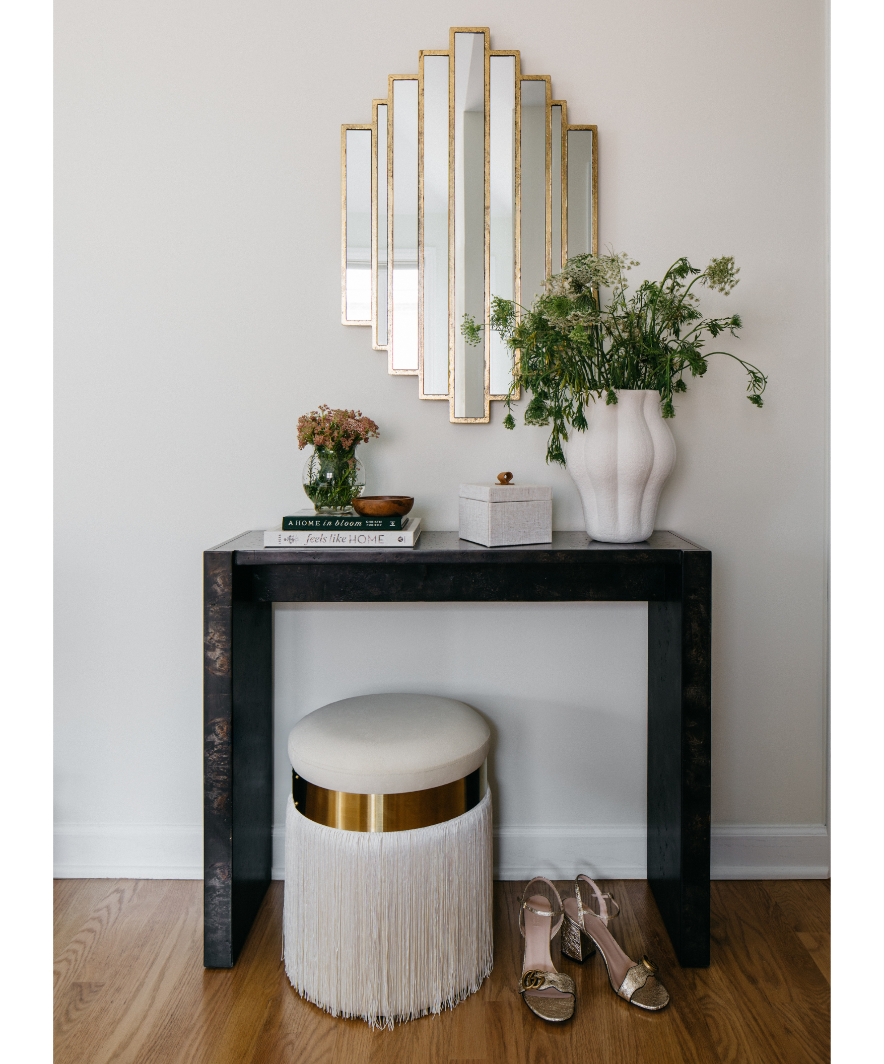 Decorative console table with books, vases with flowers, a small box, and a mirror above it, accompanied by a round cushioned stool with fringe at the bottom and a pair of open-toe heels on the wooden floor.