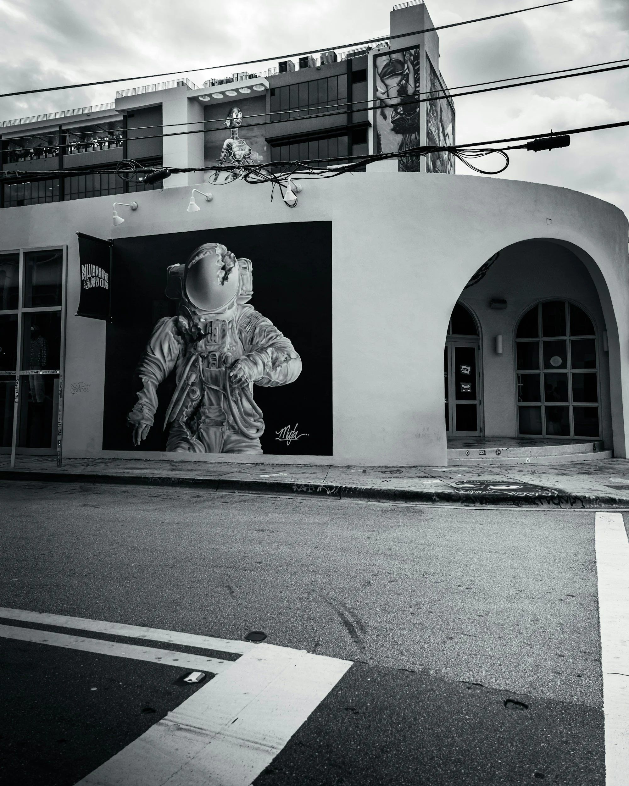 Black and white photo of a street corner with a building featuring a large mural of an astronaut in a space suit on the wall.