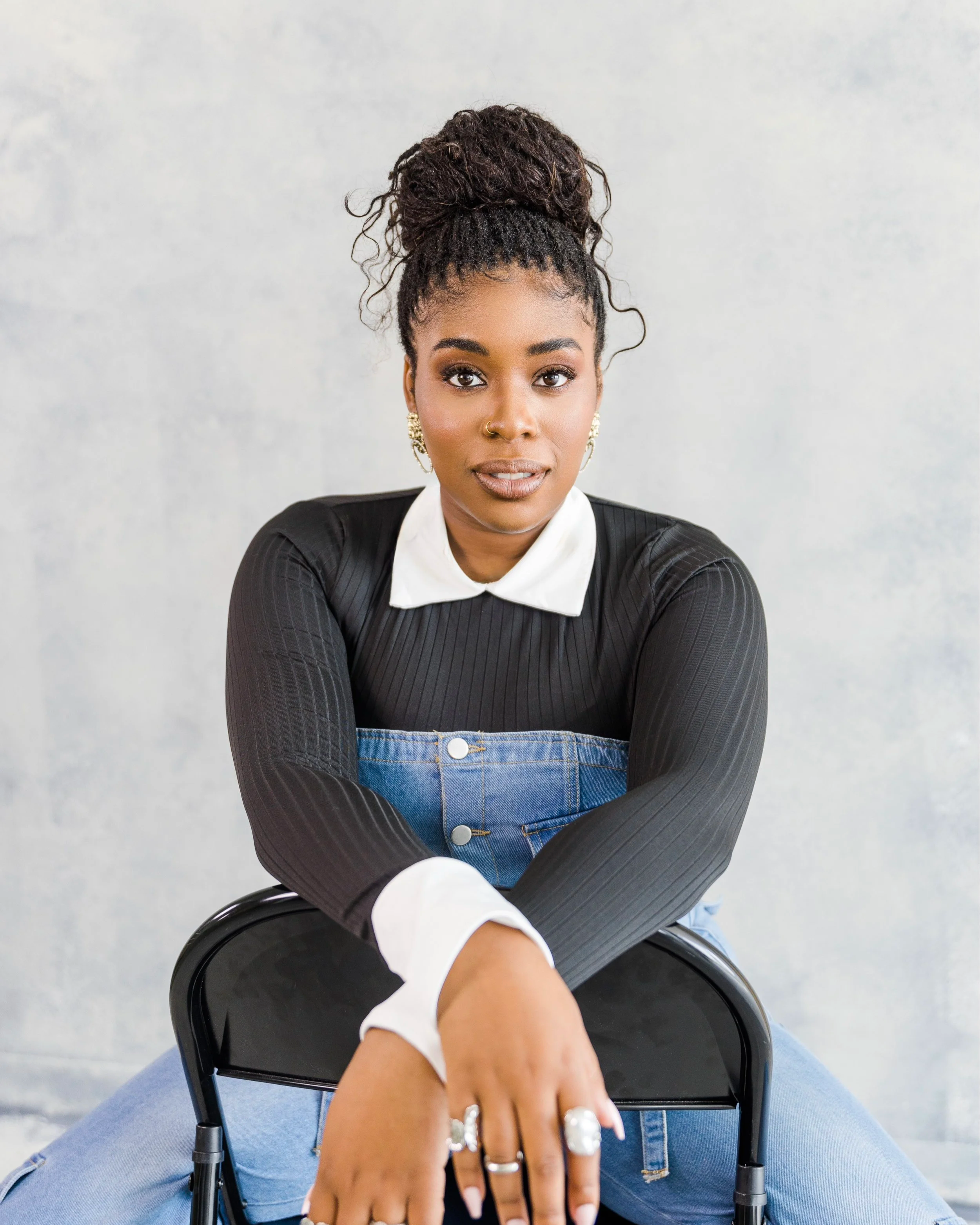 A woman with curly hair in a bun, wearing a black top with white collar and cuffs, sitting on a black chair with her knee up, looking at the camera against a gray background.