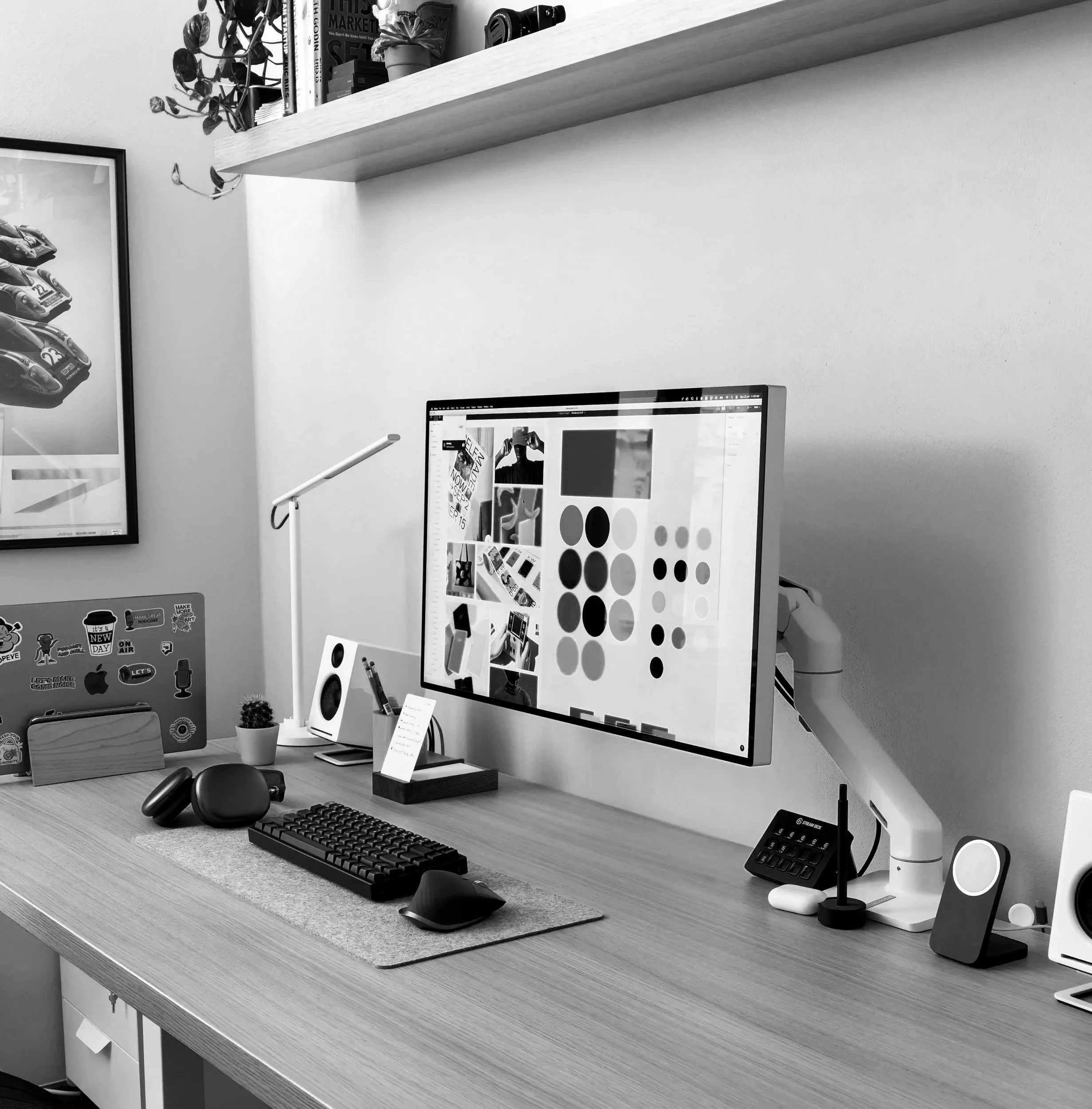 A workspace with a wooden desk, a large computer monitor displaying various color palette images, a keyboard, mouse, and speakers. There is a small potted cactus, a white speaker, and a phone stand with a phone. A white adjustable desk lamp and a printer are on the desk. A shelf with plants and books is above the workspace, and a framed poster of racing cars hangs on the wall.
