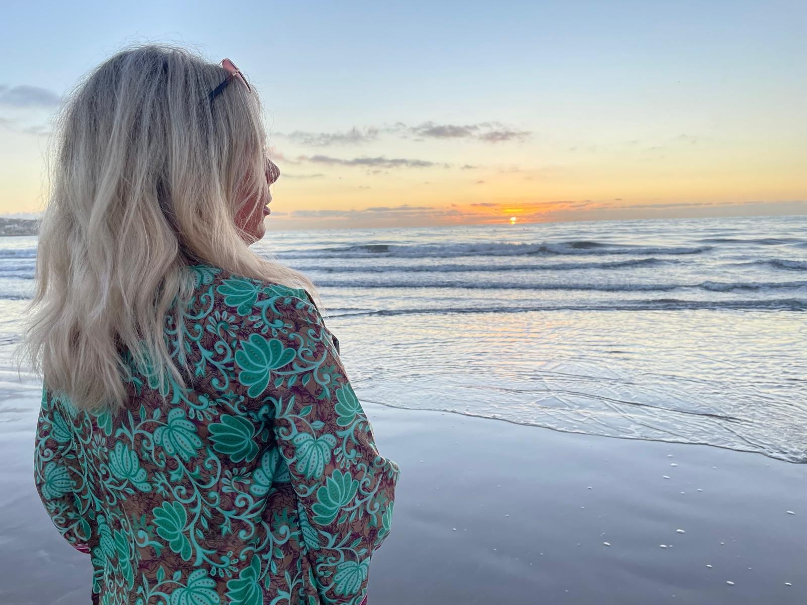 Vrouw met blond haar en bloemenpatroon jas die naar de horizon kijkt bij zonsondergang op het strand.
