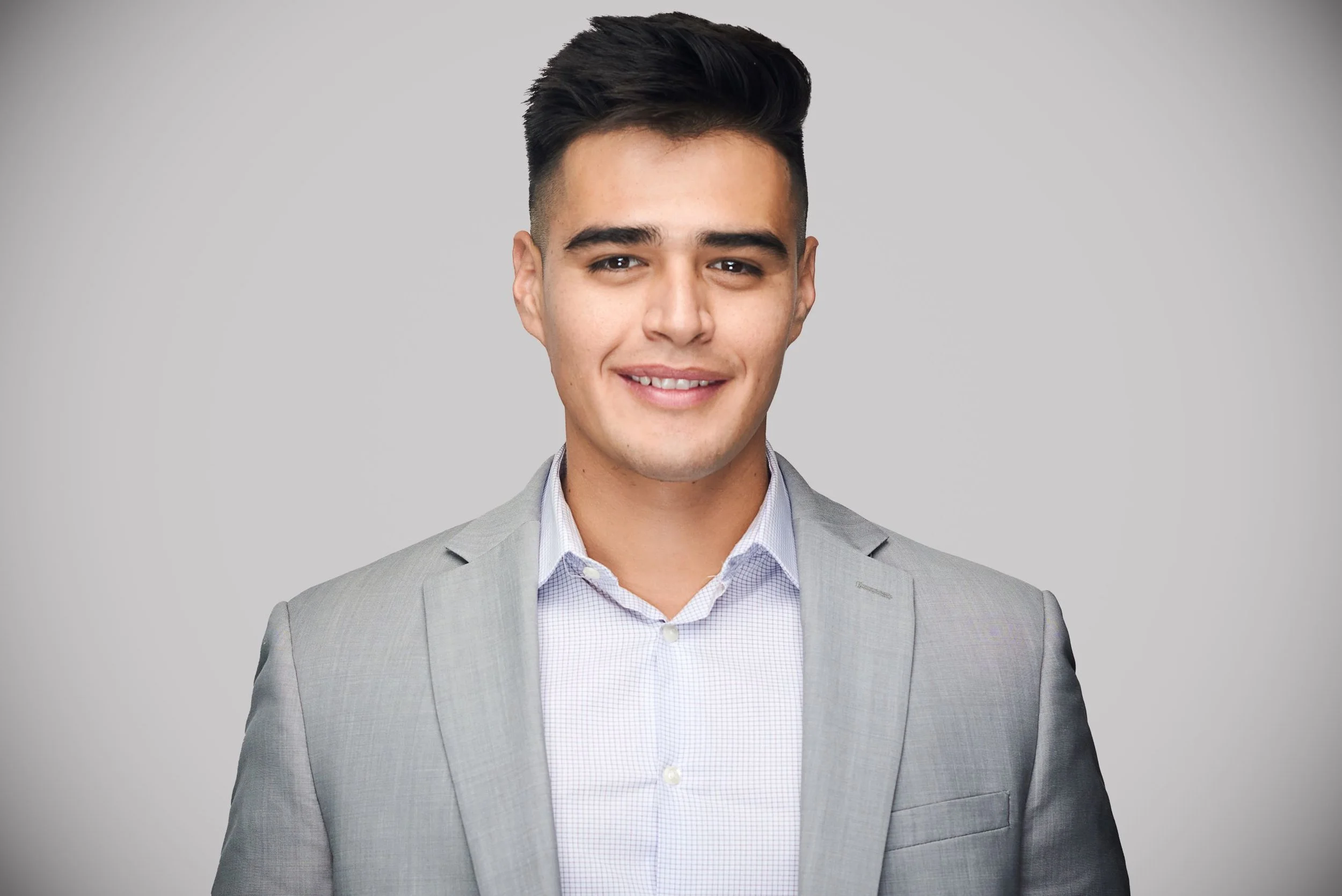 Young man in a light gray suit with a white shirt, smiling, against a plain gray background.