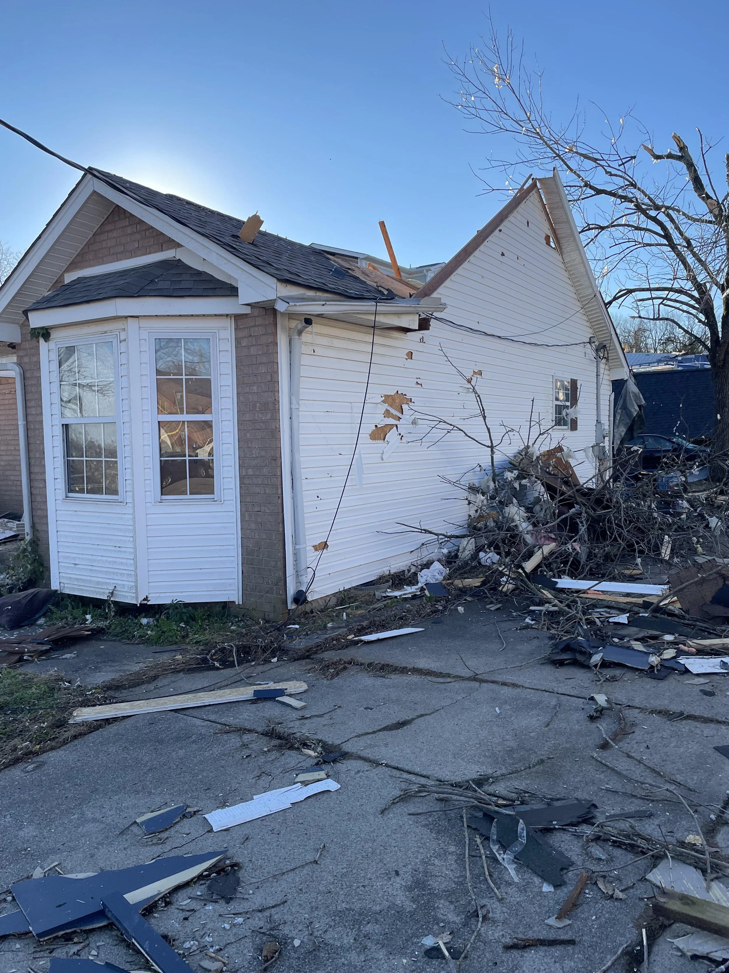 A house with significant damage from a storm or accident, with a partially collapsed roof, fallen tree branches, and debris scattered on the driveway and yard.