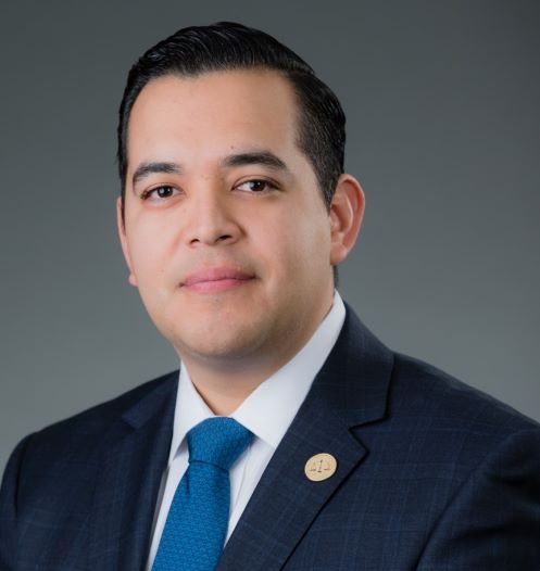 Portrait of a man in a business suit with a blue tie and a lapel pin, against a gray background.