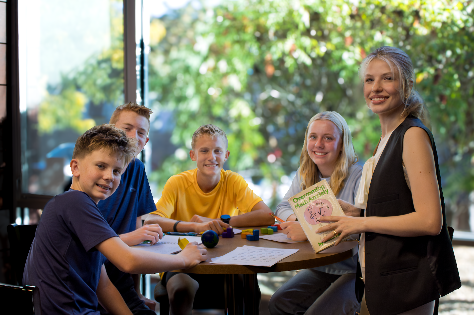 A woman and a boy sit at a round wooden table, smiling and engaging in a math learning activity with books, papers, pens, and small educational toys, with trees and sunlight in the background.