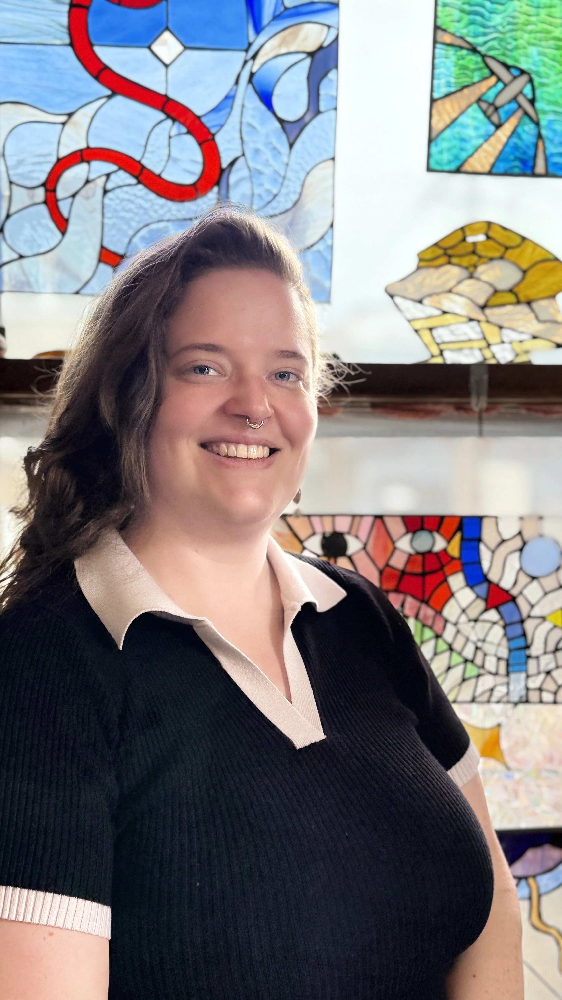 A smiling woman with long wavy brown hair, wearing a black shirt with a white collar, standing in front of colorful stained glass artwork.