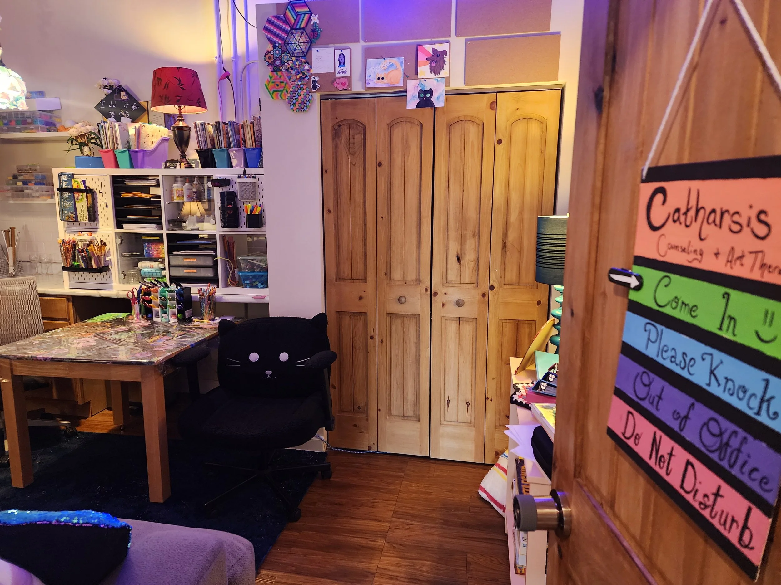 A cozy and colorful home office or craft room with wooden floors, a closed wooden closet, and a variety of art supplies and decorations. The doorway has a sign with house rules, and there is a black chair with a cute cat face cushion.