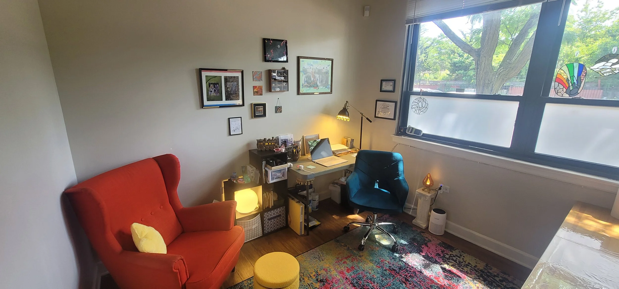 A cozy home office with a red armchair, a yellow ottoman, a patterned area rug, a black office chair, a small desk with a laptop, and framed artwork on the wall. Sunlight streams through a large window showing green trees outside.
