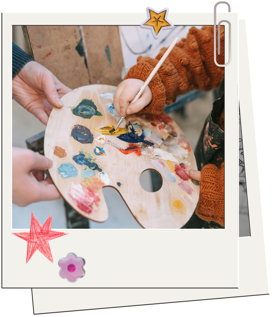 Child wearing an orange sweater painting on a wooden artist's palette with a paintbrush, assisted by an adult, in an art studio.