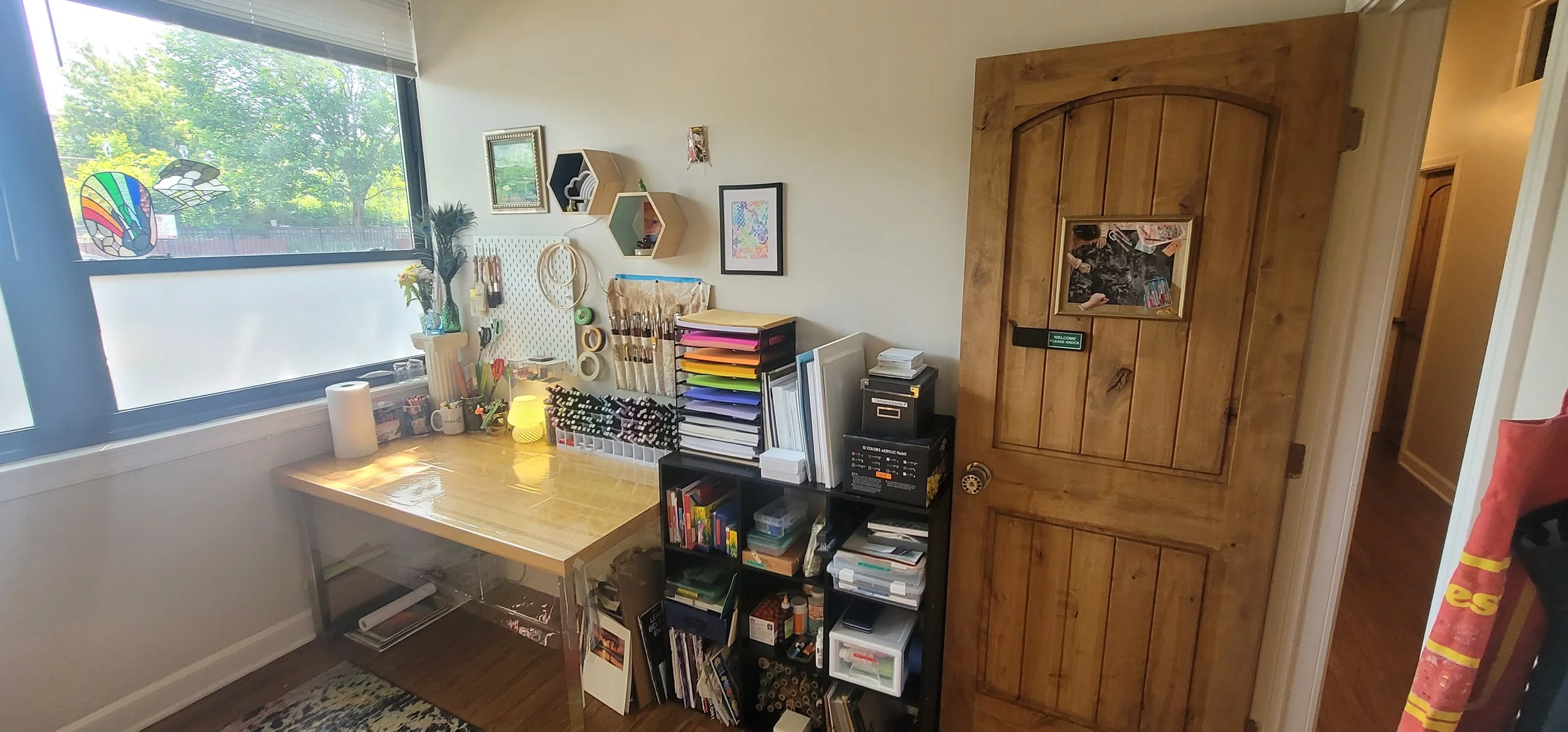 Home office with wooden door, organized desk with art supplies, shelves, and window with stained glass