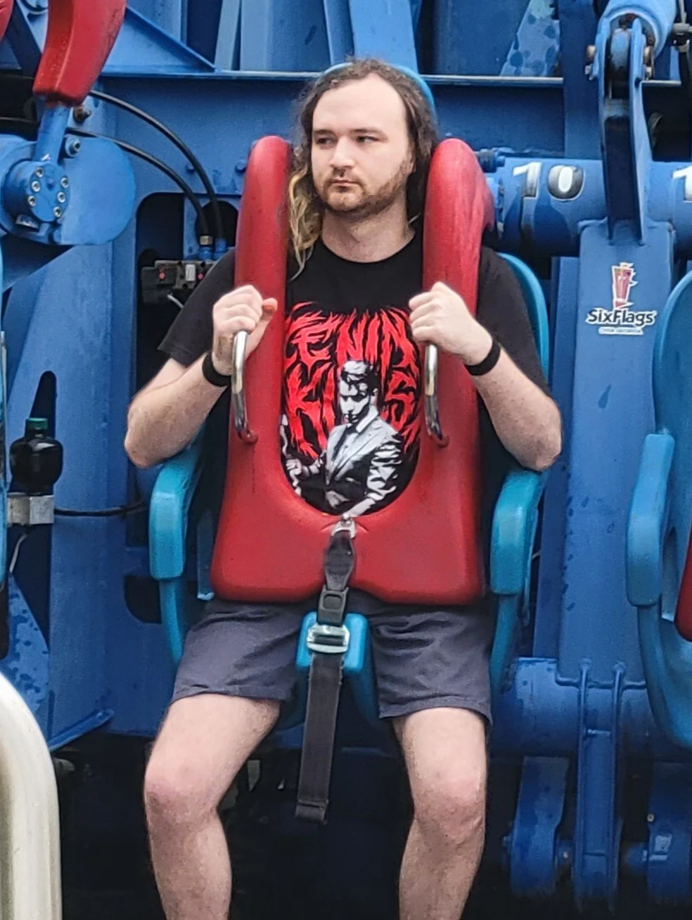 A man with long, curly hair and a beard seated on a thrill ride at Six Flags amusement park, appearing to be scared, wearing a black T-shirt with a red and white graphic design and shorts.