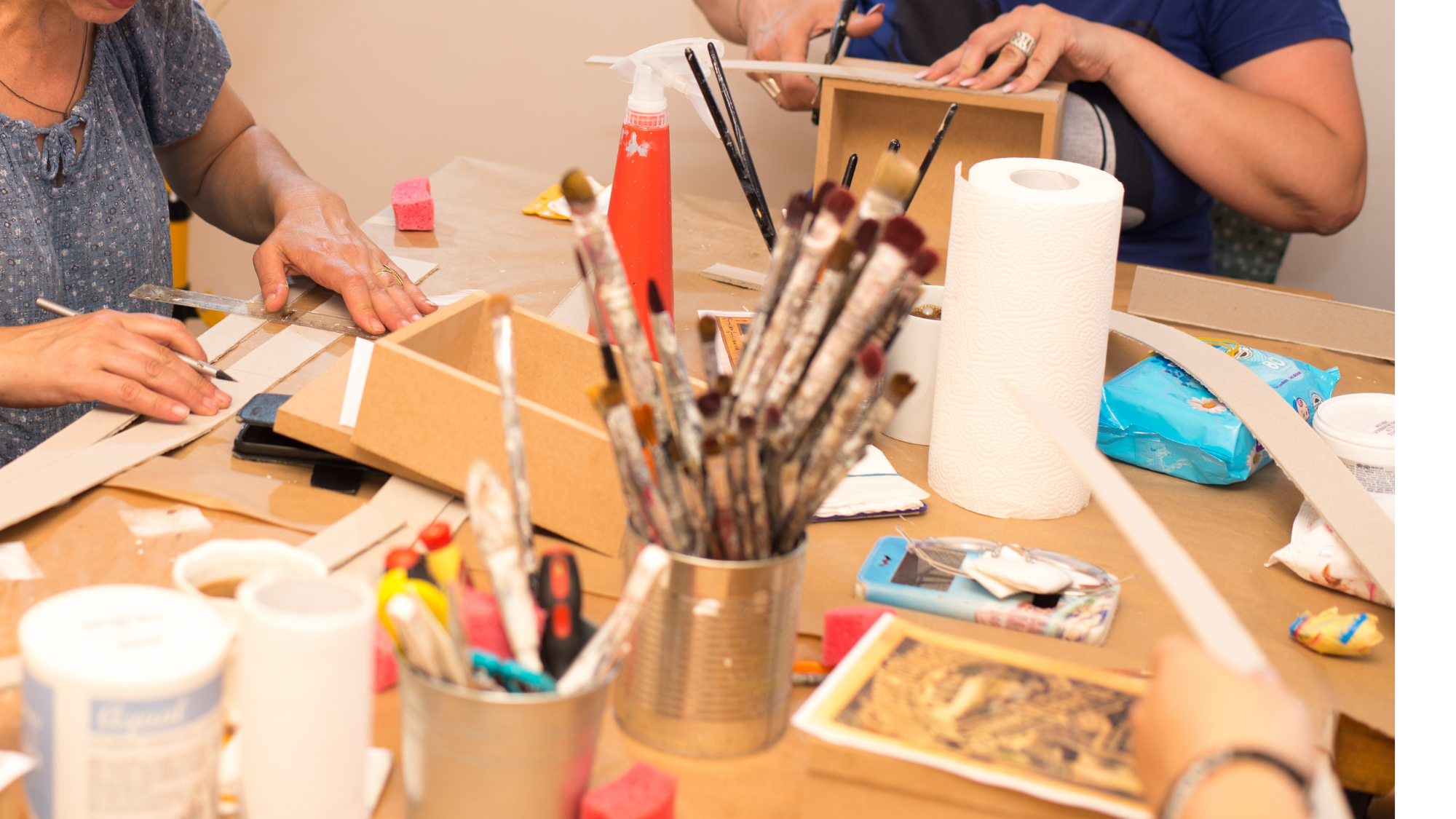People working on a craft project at a cluttered table with brushes, paper towels, and art supplies.
