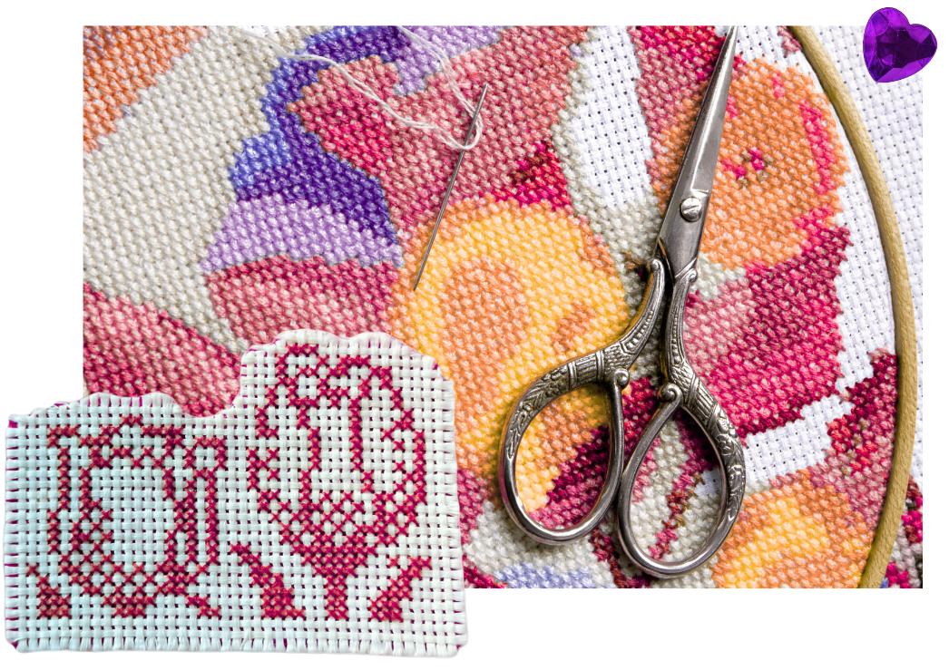 Embroidery hoop with a floral cross-stitch design, embroidery scissors, sewing needle with pink thread, and a piece of white cross-stitch fabric with a red design, all on a colorful fabric background.