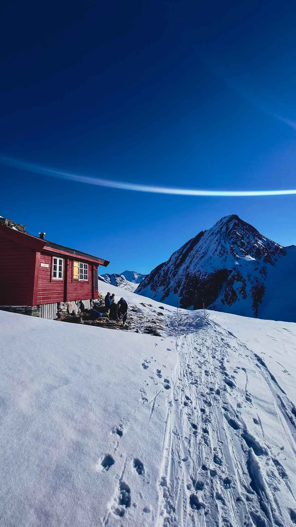 Snøbelagt fjellandskap med en rød hytte, tre personer og spor i snøen under klar blå himmel.