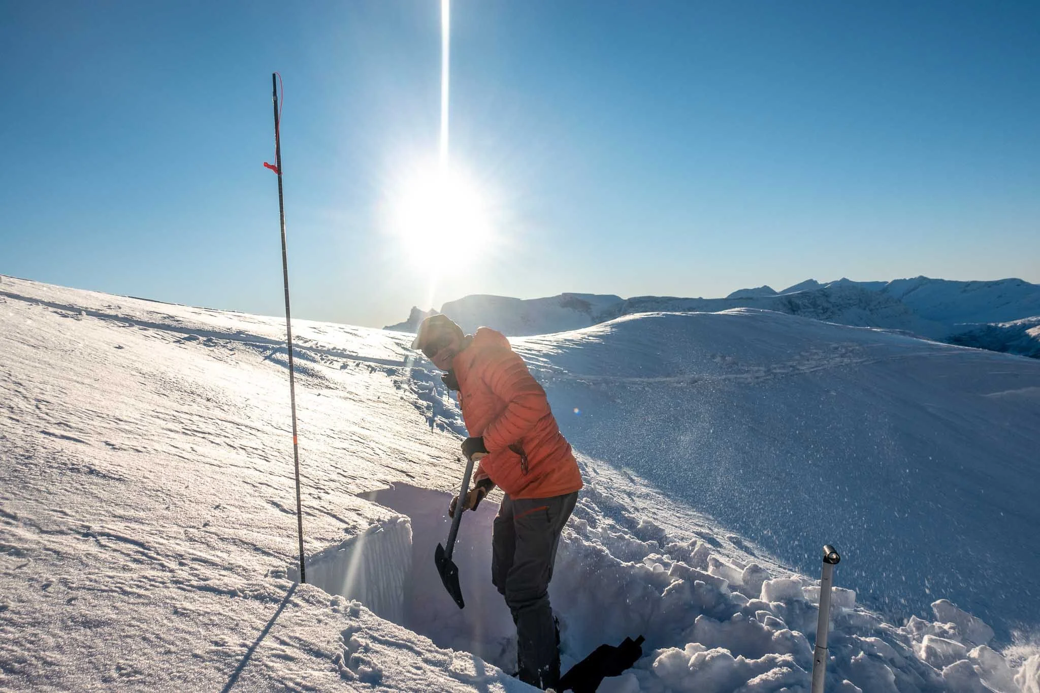 En person i oransje jakke og svarte bukser graver i snø med en spade på en snødekt fjelltopp under klar blå himmel.