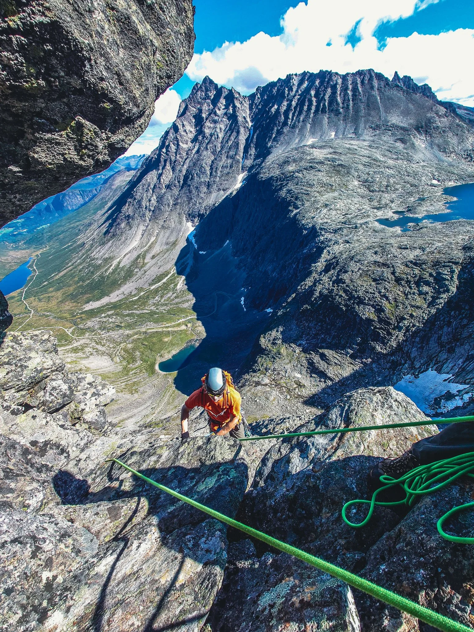 En fjellklatrer i orange utstyr klatrer opp en bratt fjellvegg med tau og sikringer, omgitt av høye fjell og en klar blå himmel.