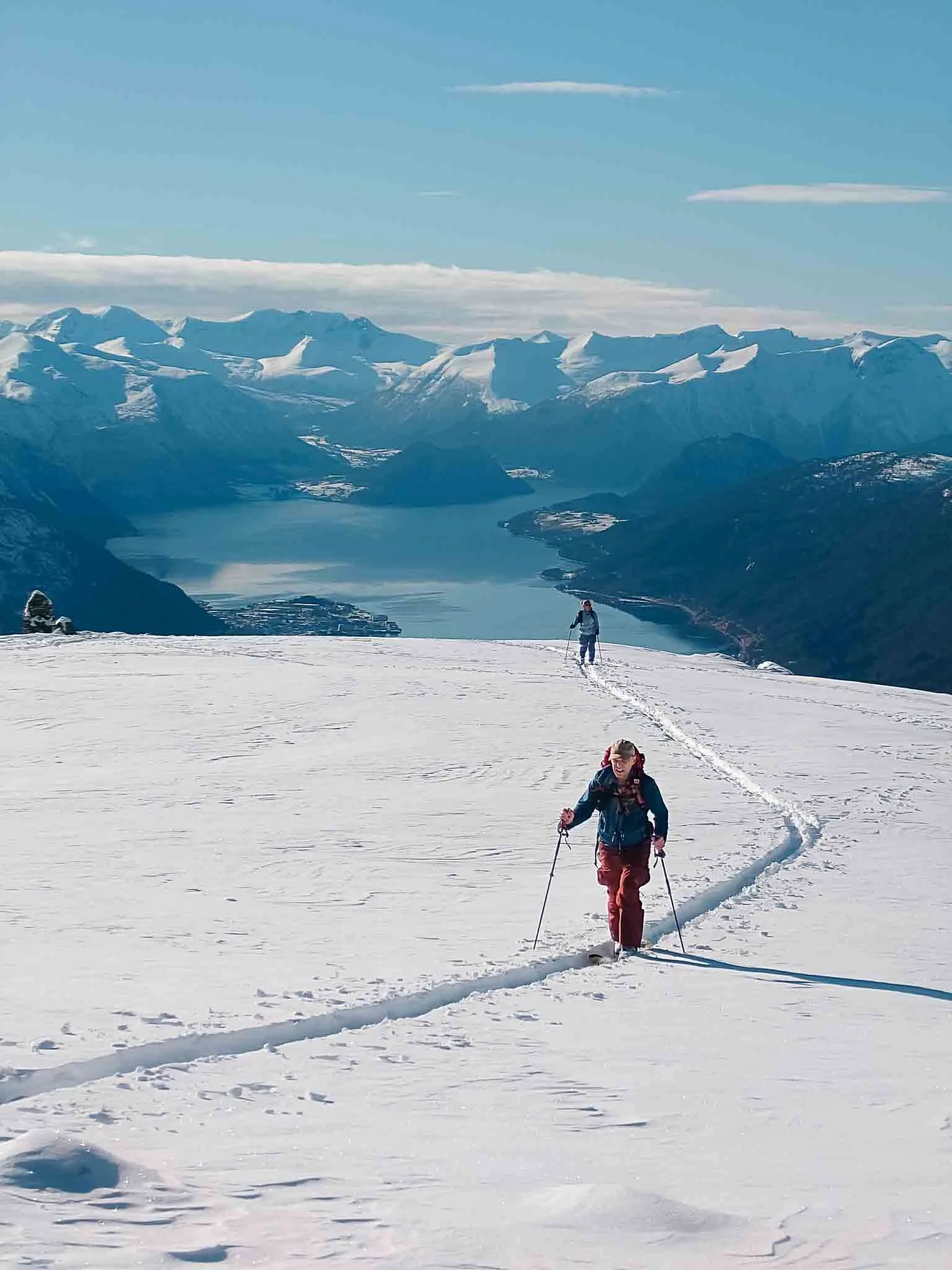 To personer går på snø med ski, omgitt av fjell og fjord, under en klar blå himmel.