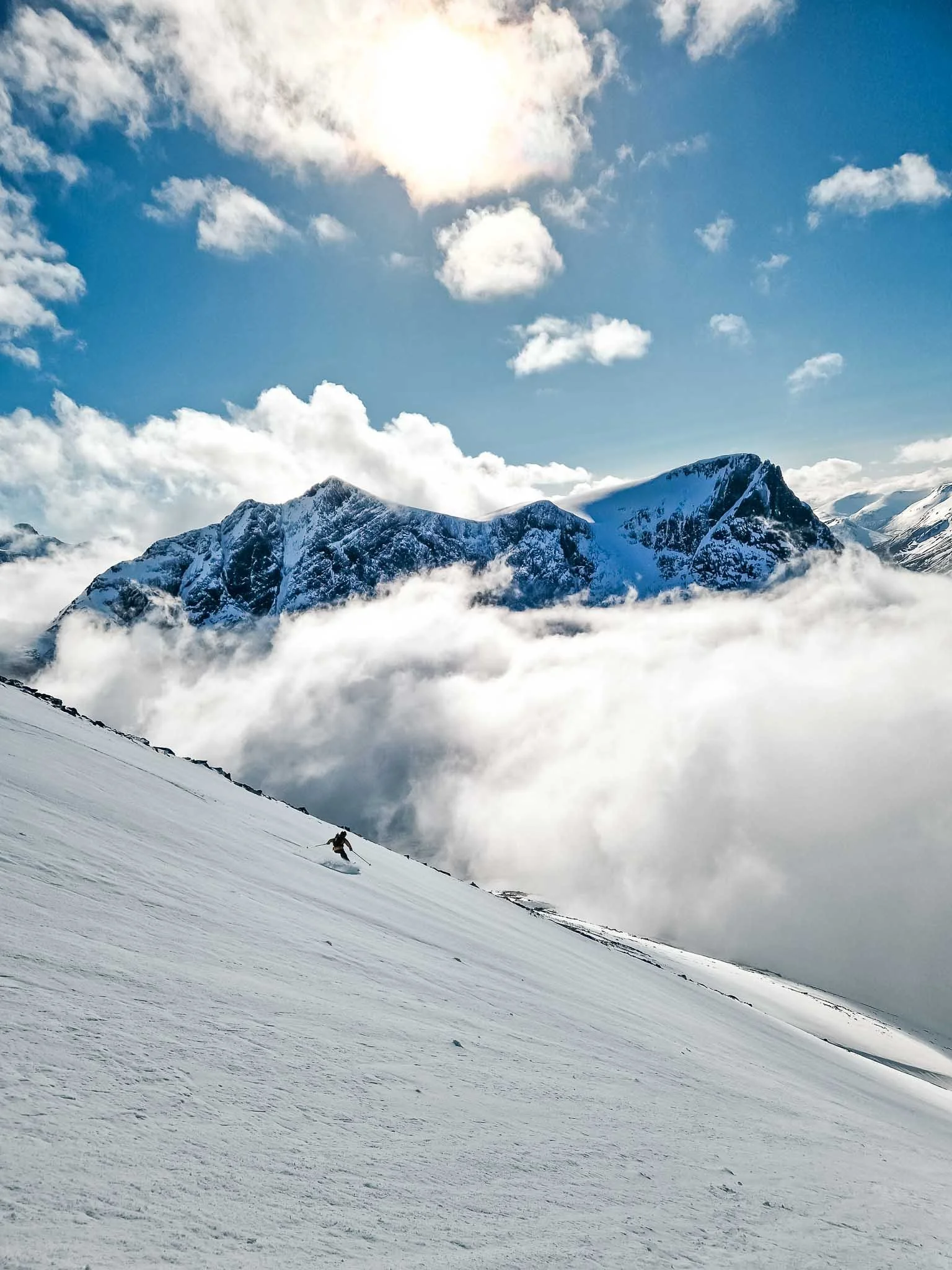 En skisport utøver kjører ned en snødekt fjellside med fjell og skyer i bakgrunnen under en solrik dag.