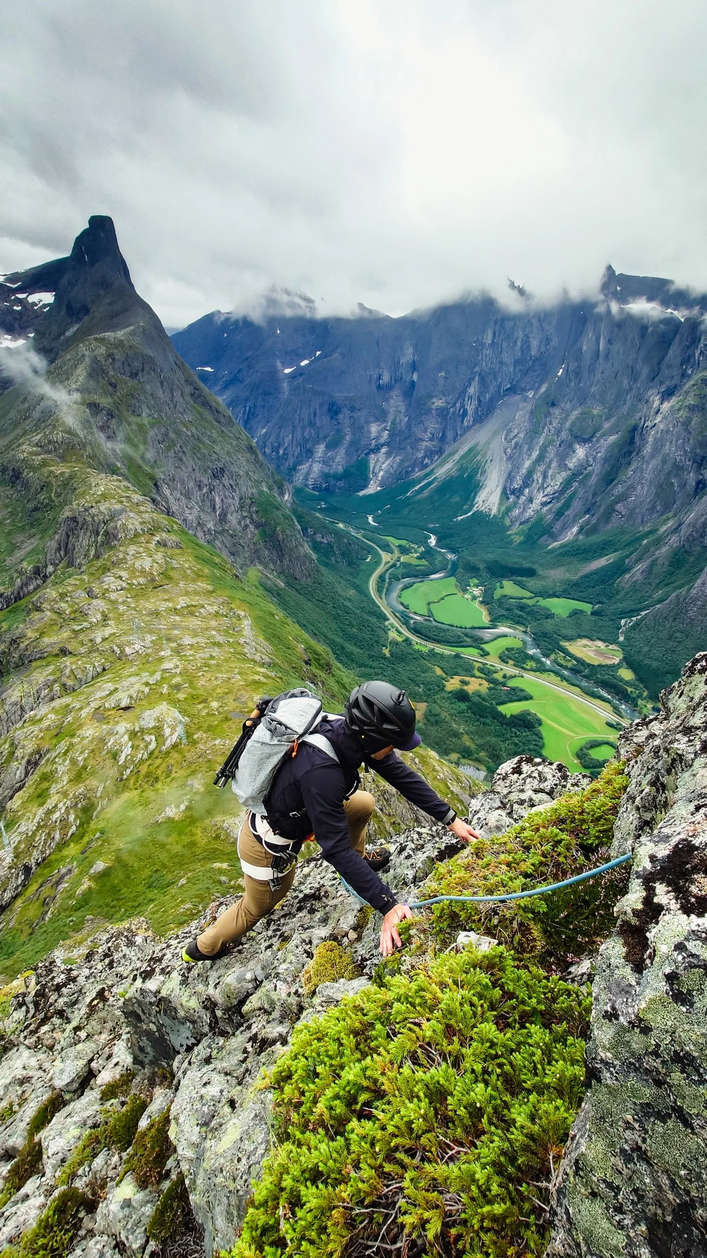 En person klatrer oppover en fjellside med fjell i bakgrunnen og en dal med elv, enger og skog nedenfor.