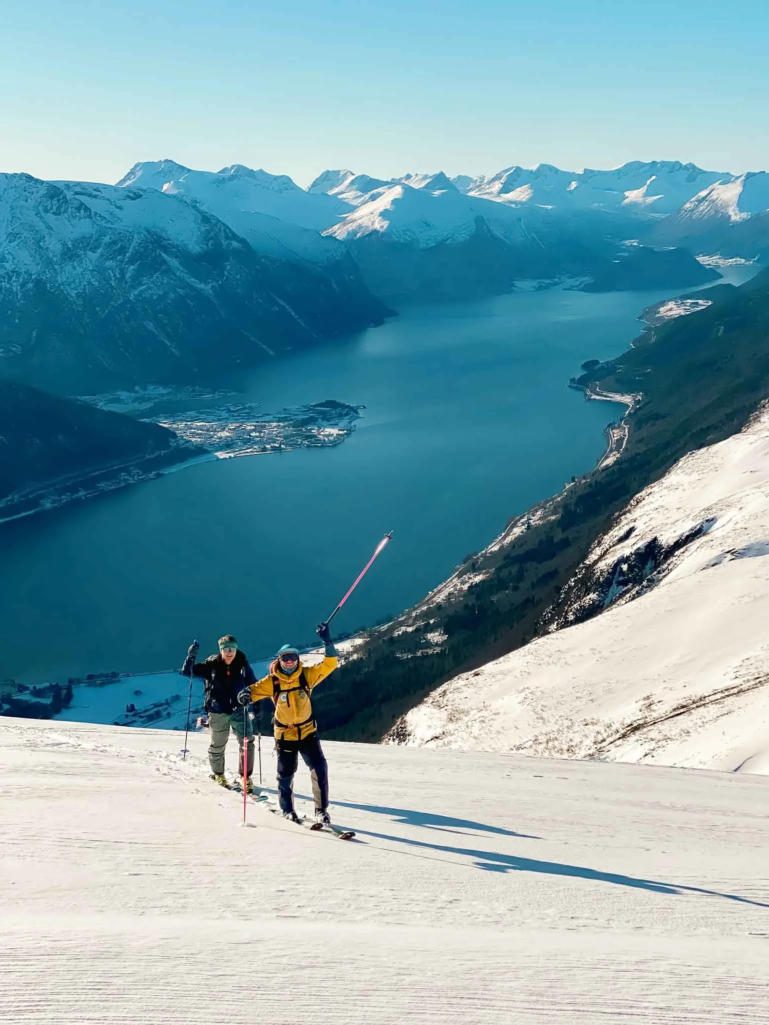 To personer paraglider nedover en snødekt fjelltopp med innsjø og fjell i bakgrunnen.